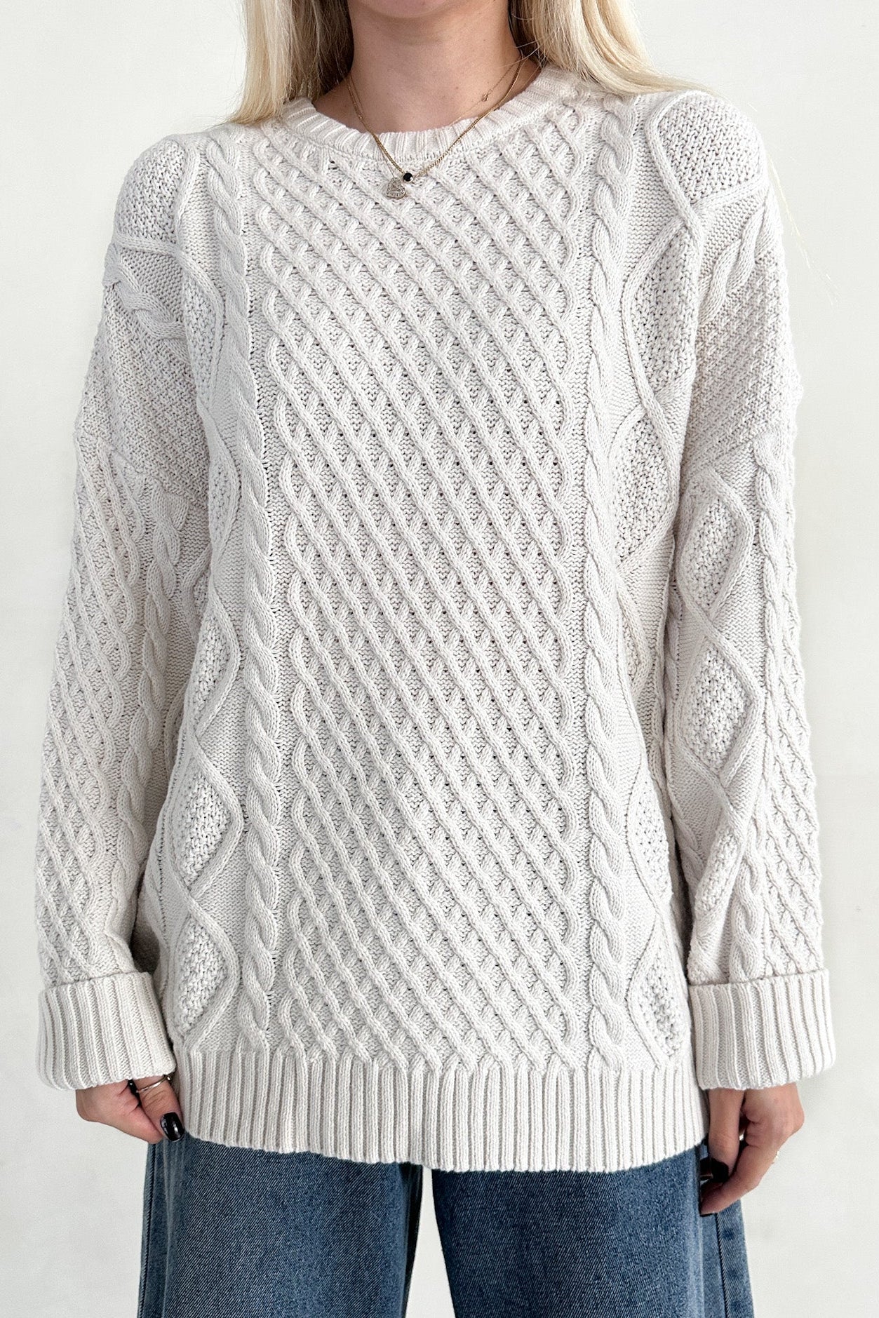 Person wearing a white cable knit sweater and blue jeans on a plain background