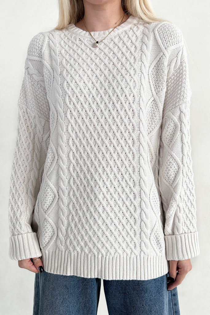 Person wearing a white cable knit sweater and blue jeans on a plain background