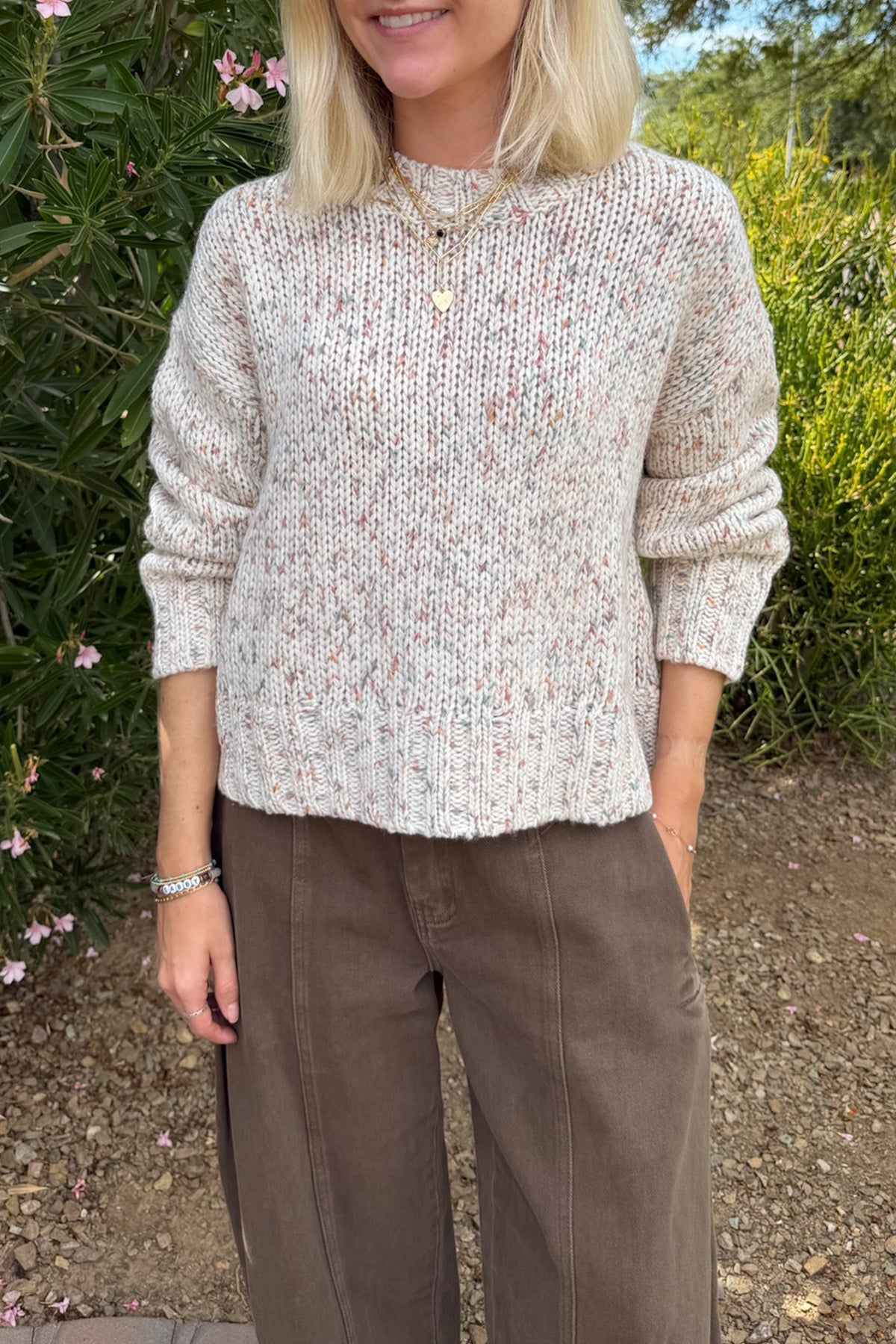A woman wearing a cream-colored, textured knit sweater with subtle multicolor flecks, paired with brown wide-leg trousers, standing outdoors in front of green foliage.