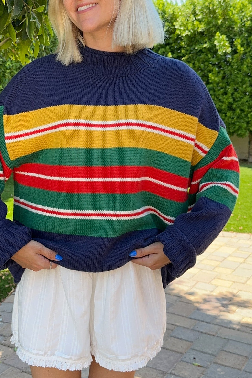 A woman wearing a colorful striped knit sweater with navy blue, yellow, green, red, and white horizontal stripes, paired with white shorts and tan boots, standing outdoors on a paved walkway with green hedges in the background.