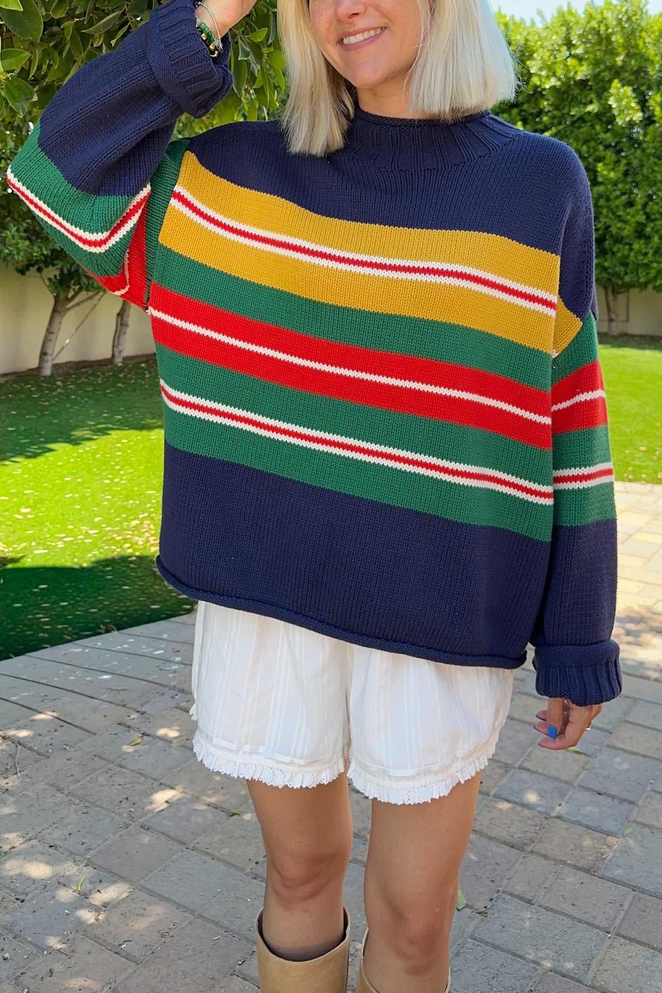 A woman wearing a navy blue knit sweater with bold horizontal stripes in yellow, green, red, and white, paired with white shorts, standing outdoors on a paved walkway with green grass in the background.