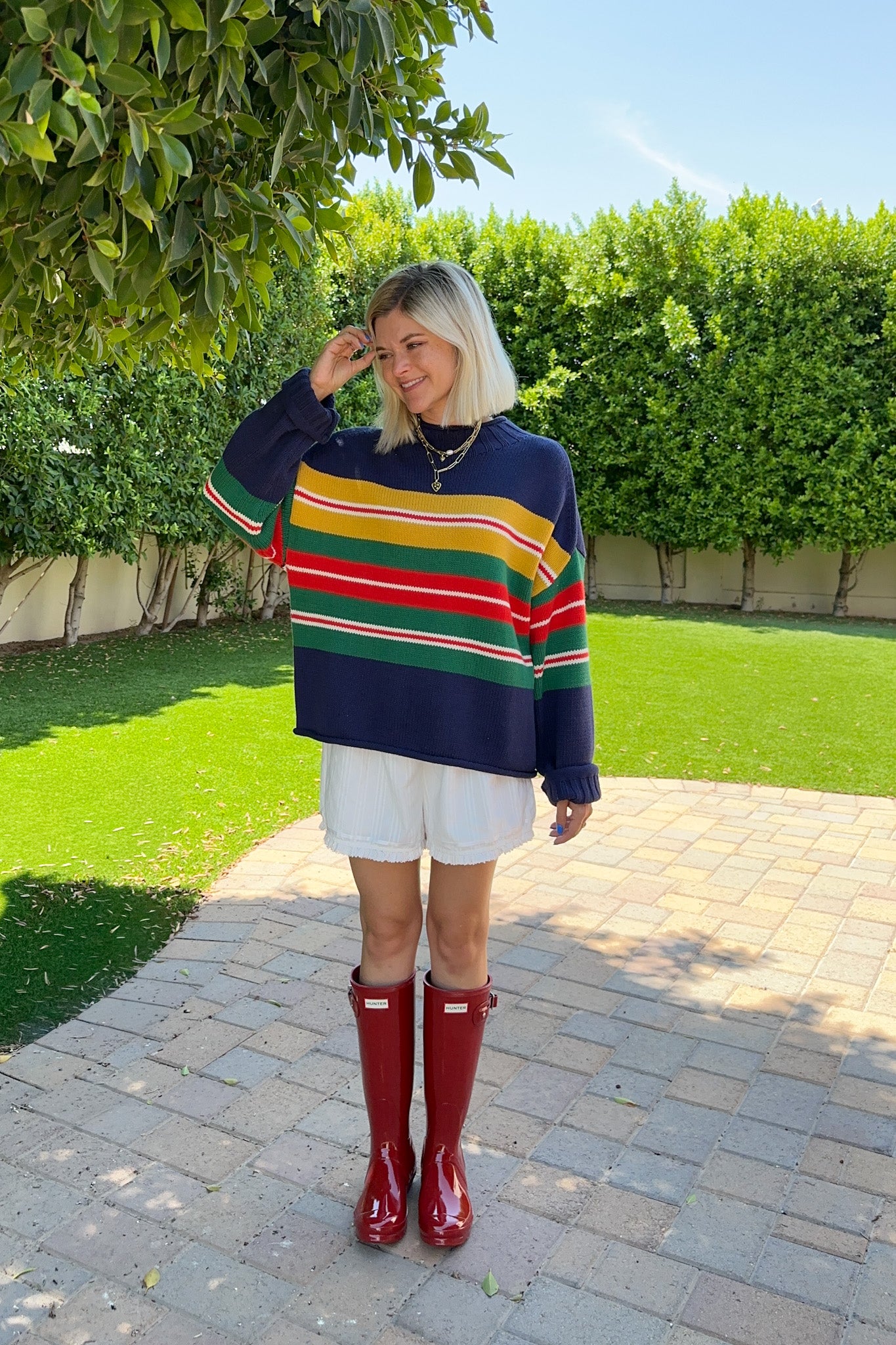 A woman stands outdoors on a paved path in a garden, wearing a navy blue knit sweater with horizontal stripes of yellow, green, red, and white, paired with white shorts and shiny red rain boots.