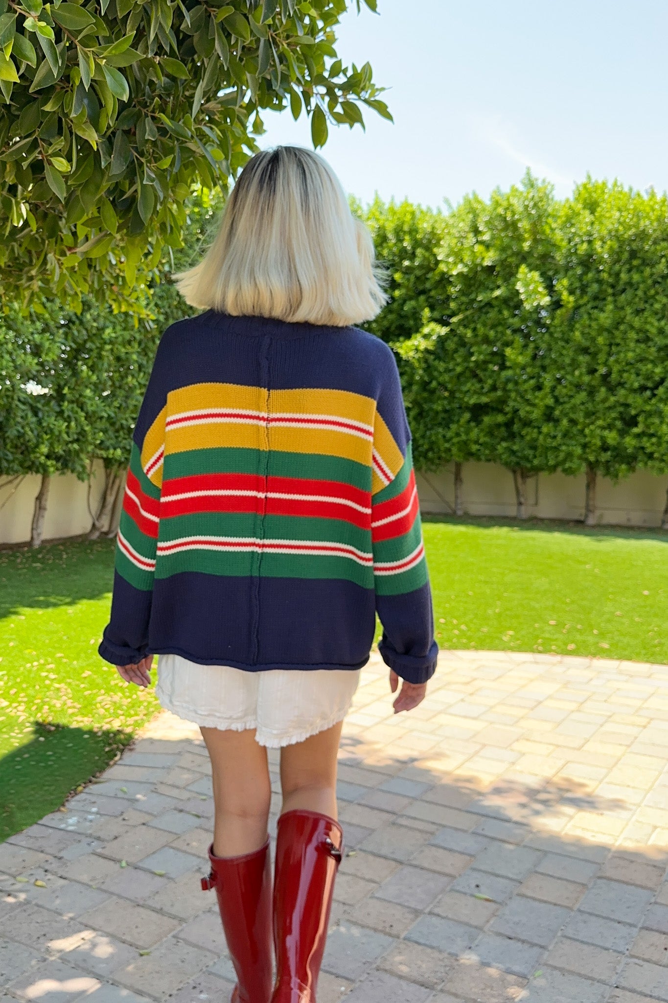 A person viewed from behind wearing a navy blue knit sweater with bold horizontal stripes in yellow, red, green, and white, paired with a white skirt and red rain boots, standing on a paved path in a garden.