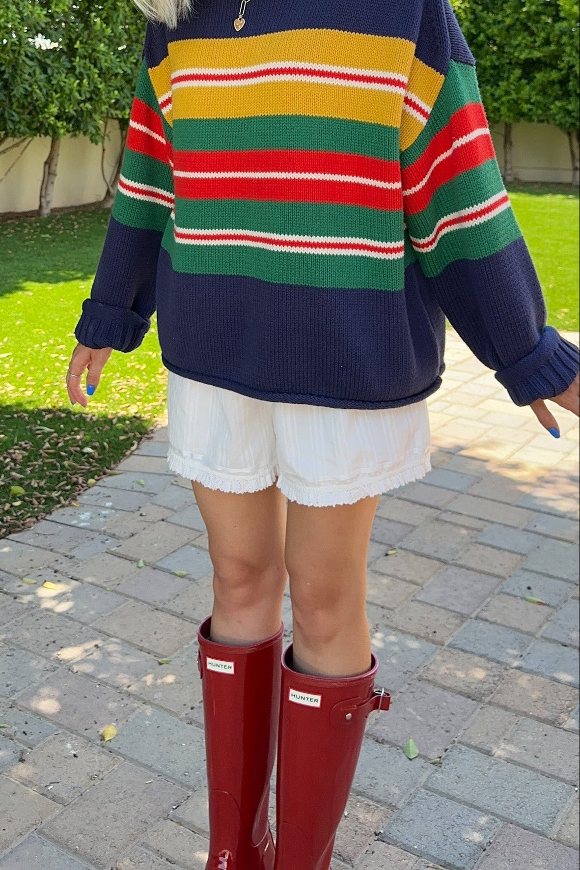 A woman wearing an oversized navy sweater with colorful horizontal stripes in yellow, green, and red, paired with white shorts and red rain boots, standing outdoors on a paved walkway with grass and trees in the background.