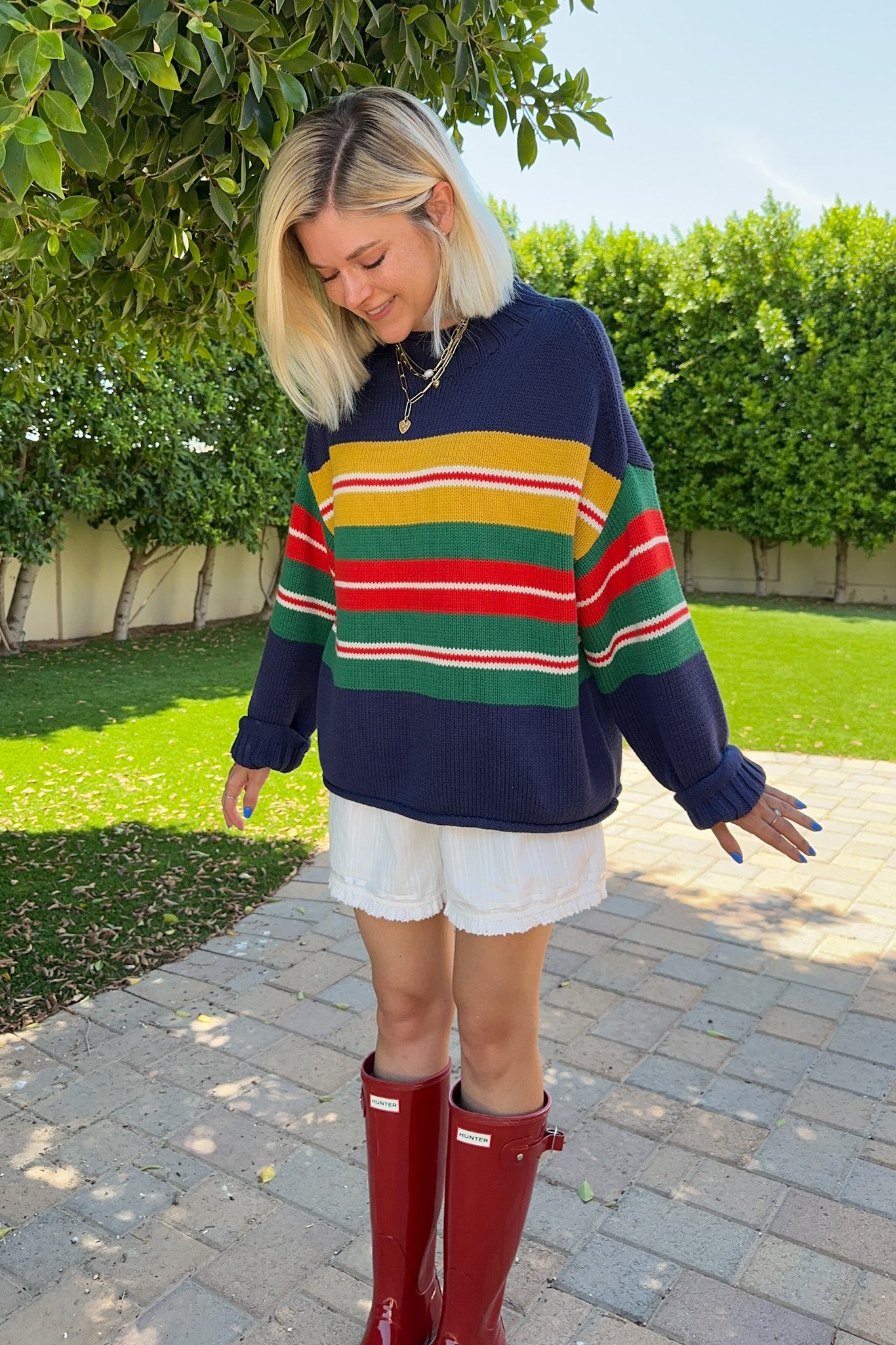 A woman wearing a navy blue knit sweater with colorful horizontal stripes in yellow, red, and green, paired with white shorts and red rain boots, standing outdoors on a paved walkway with green hedges in the background.