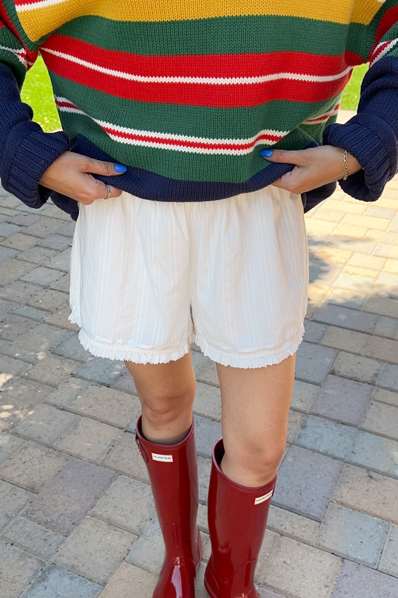 A person outdoors wearing a colorful striped knit sweater, white shorts, and red rain boots on a paved walkway.
