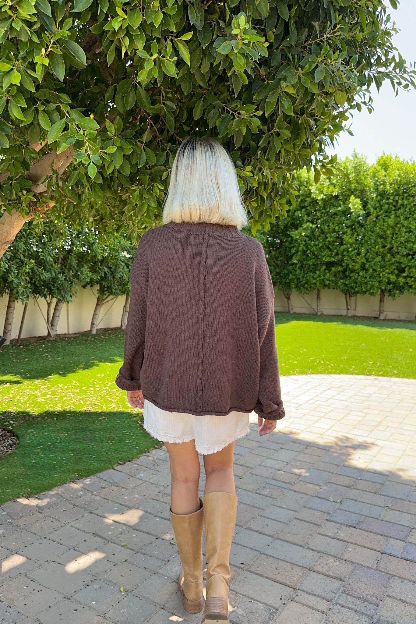 A woman viewed from behind wearing a brown knit sweater, a white ruffled skirt, and tan knee-high boots, standing on a paved path in a garden with green trees and hedges.