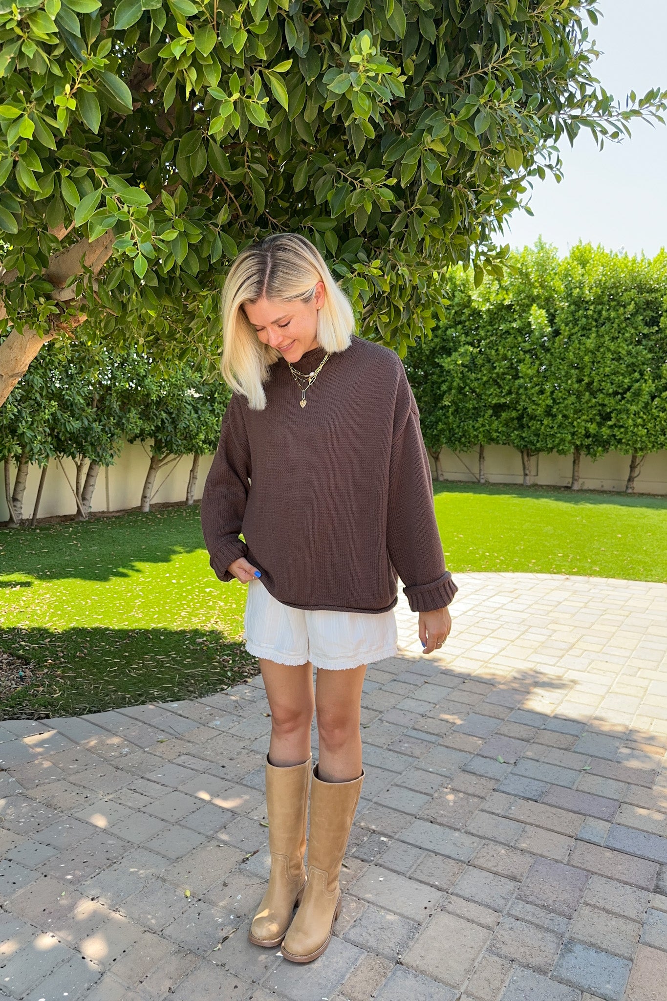 A woman standing outdoors on a paved walkway in a garden, wearing a brown ribbed mock-neck long-sleeve sweater, white shorts, and tan knee-high boots.