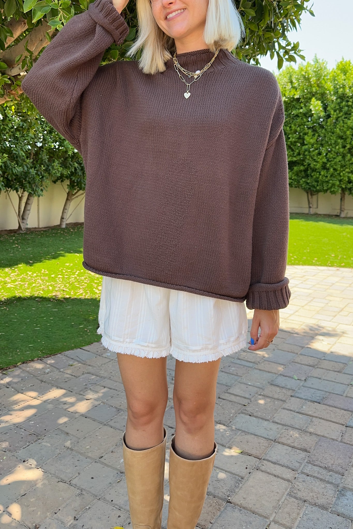 A woman wearing a brown ribbed knit mock neck sweater, white shorts, and tan boots, standing outdoors on a paved walkway with green grass and trees in the background.