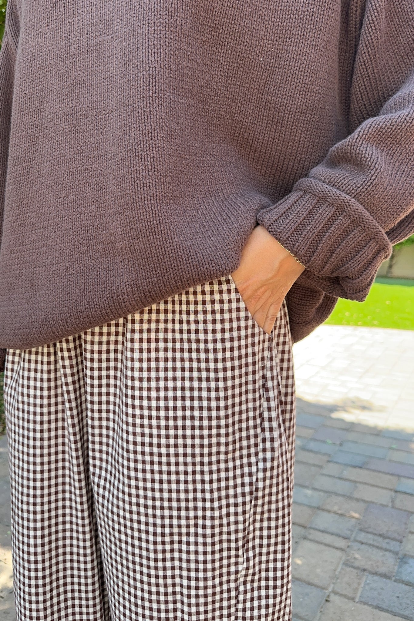 A person wearing a taupe knit sweater and brown-and-white gingham wide-leg pants, standing outdoors on a paved path with grass in the background.
