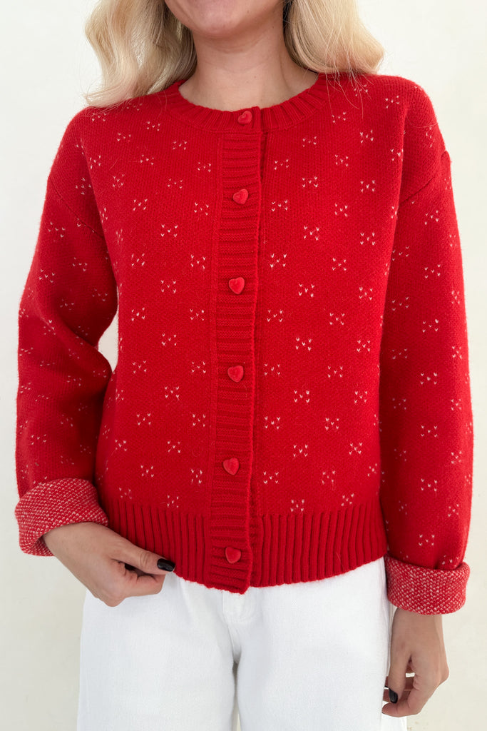 Red knitted cardigan with heart patterns worn by a person on a white background