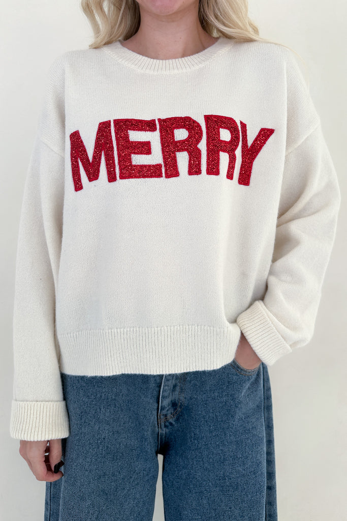 Person wearing a cream sweater with 'MERRY' in red text, standing against a plain background.