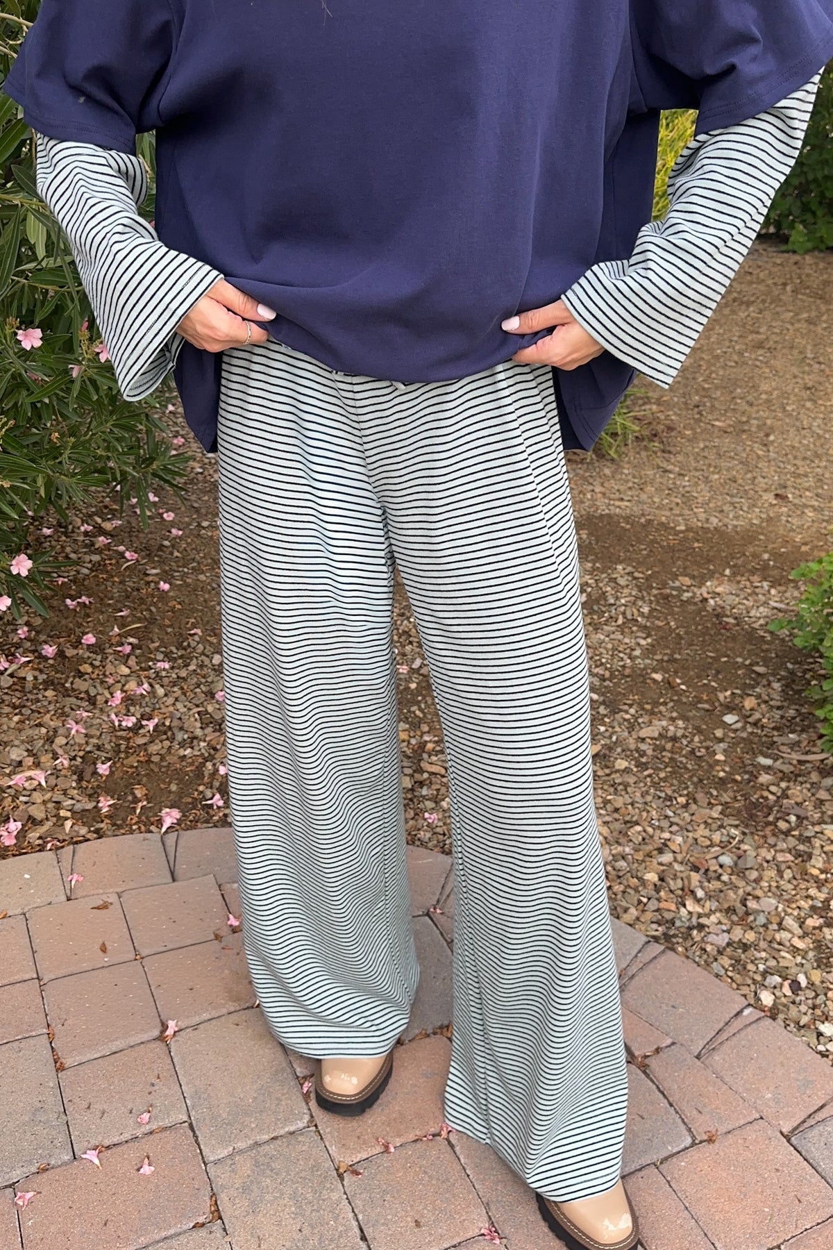A person wearing wide-leg striped pants in a garden setting. The pants feature thin black and white horizontal stripes and a relaxed fit, paired with a navy blue top and brown shoes.