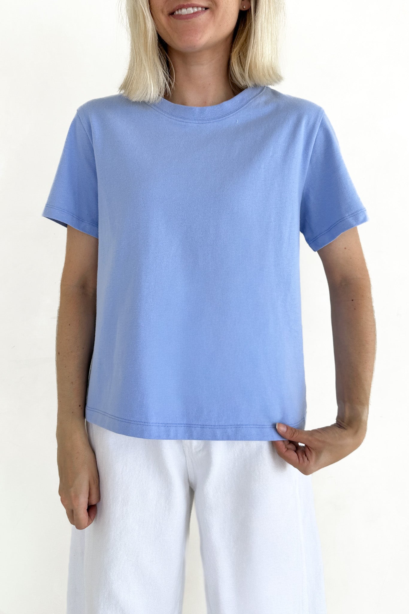 A woman wearing a light blue short-sleeve crewneck t-shirt and white pants, shown from the chin down against a plain white background.