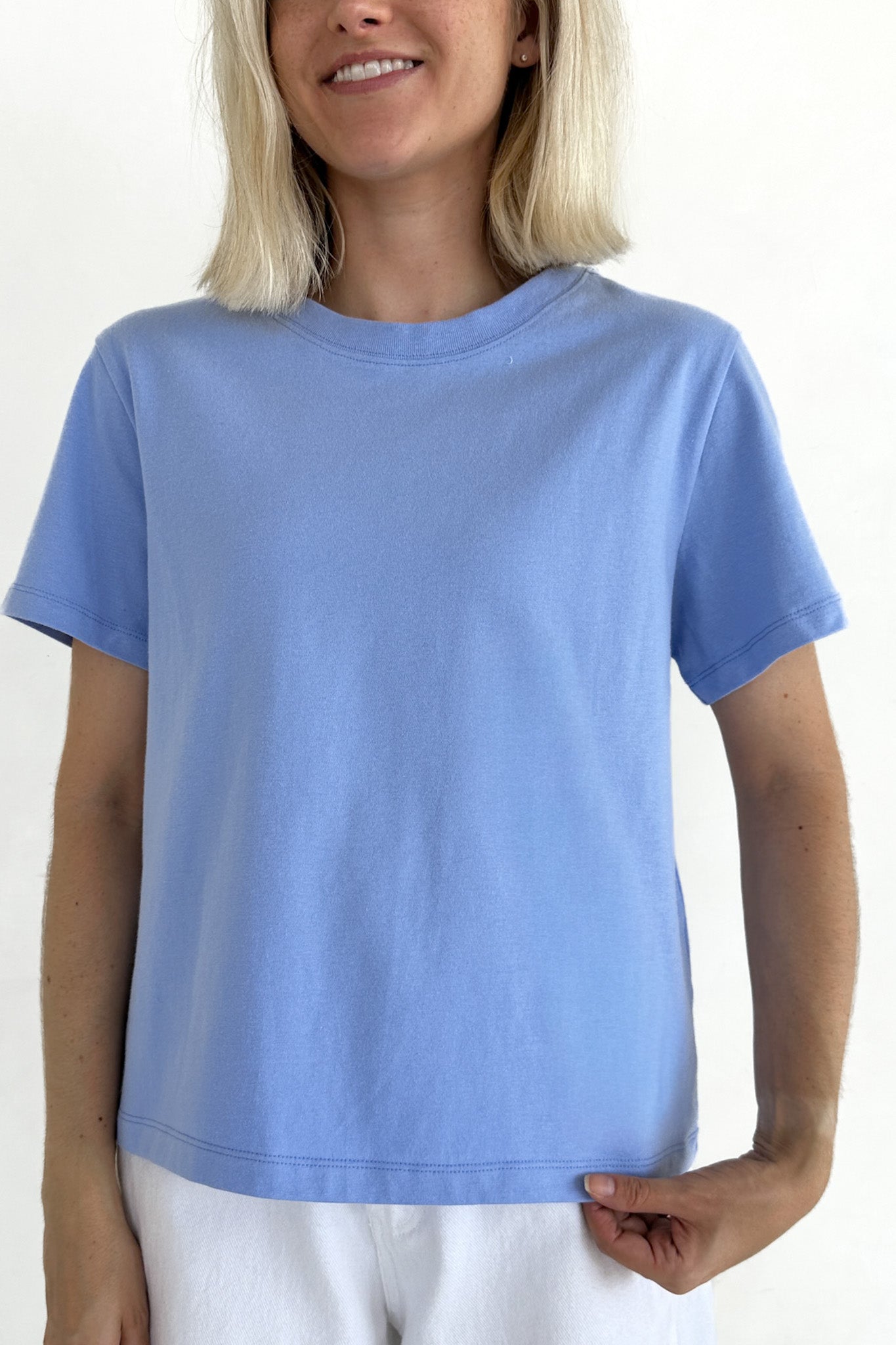 A woman wearing a light blue short-sleeve crewneck t-shirt and white pants, shown from the waist up against a plain white background.