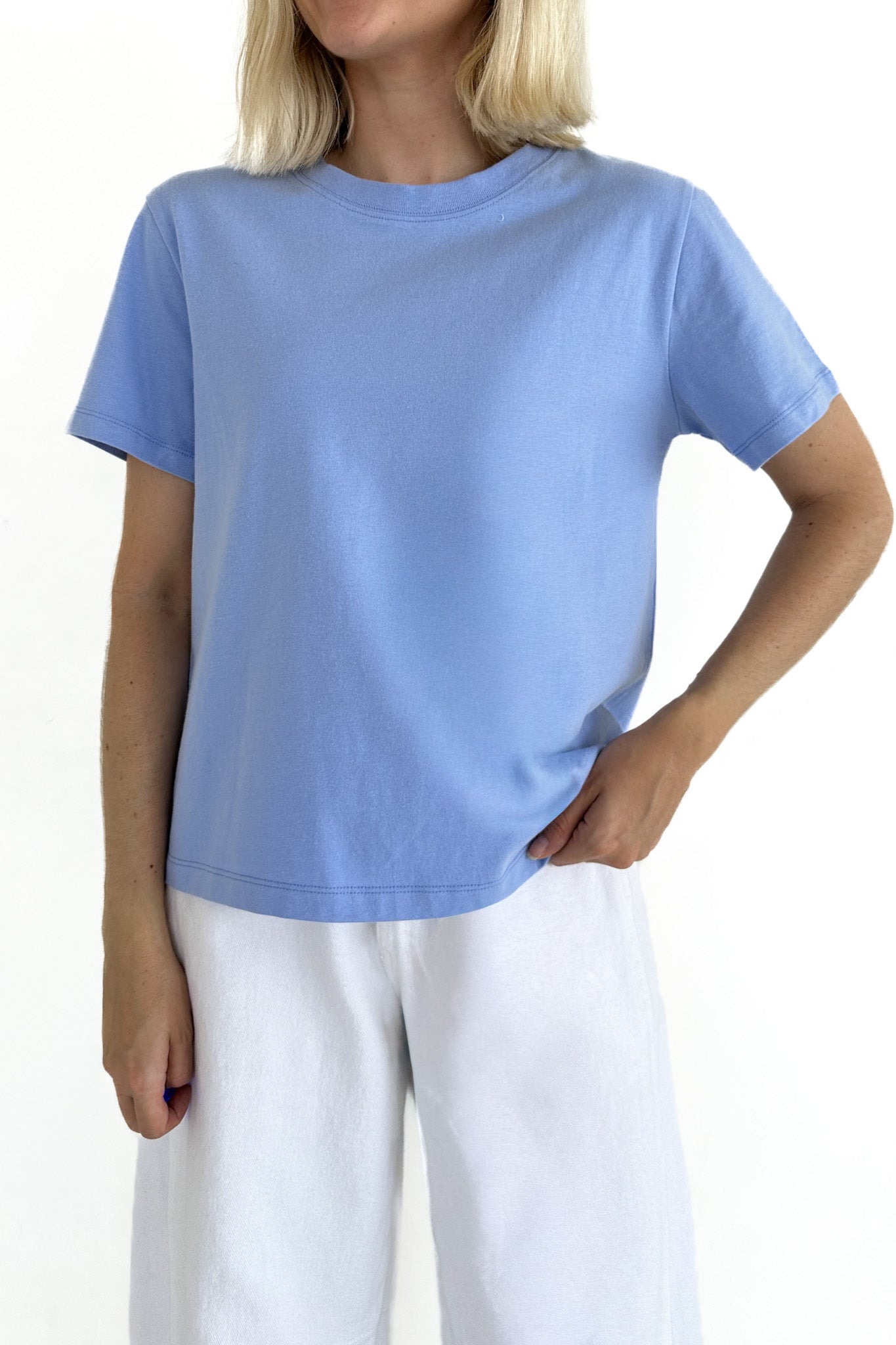 A woman wearing a light blue short-sleeve crewneck t-shirt and white pants, shown from the neck down against a plain white background.