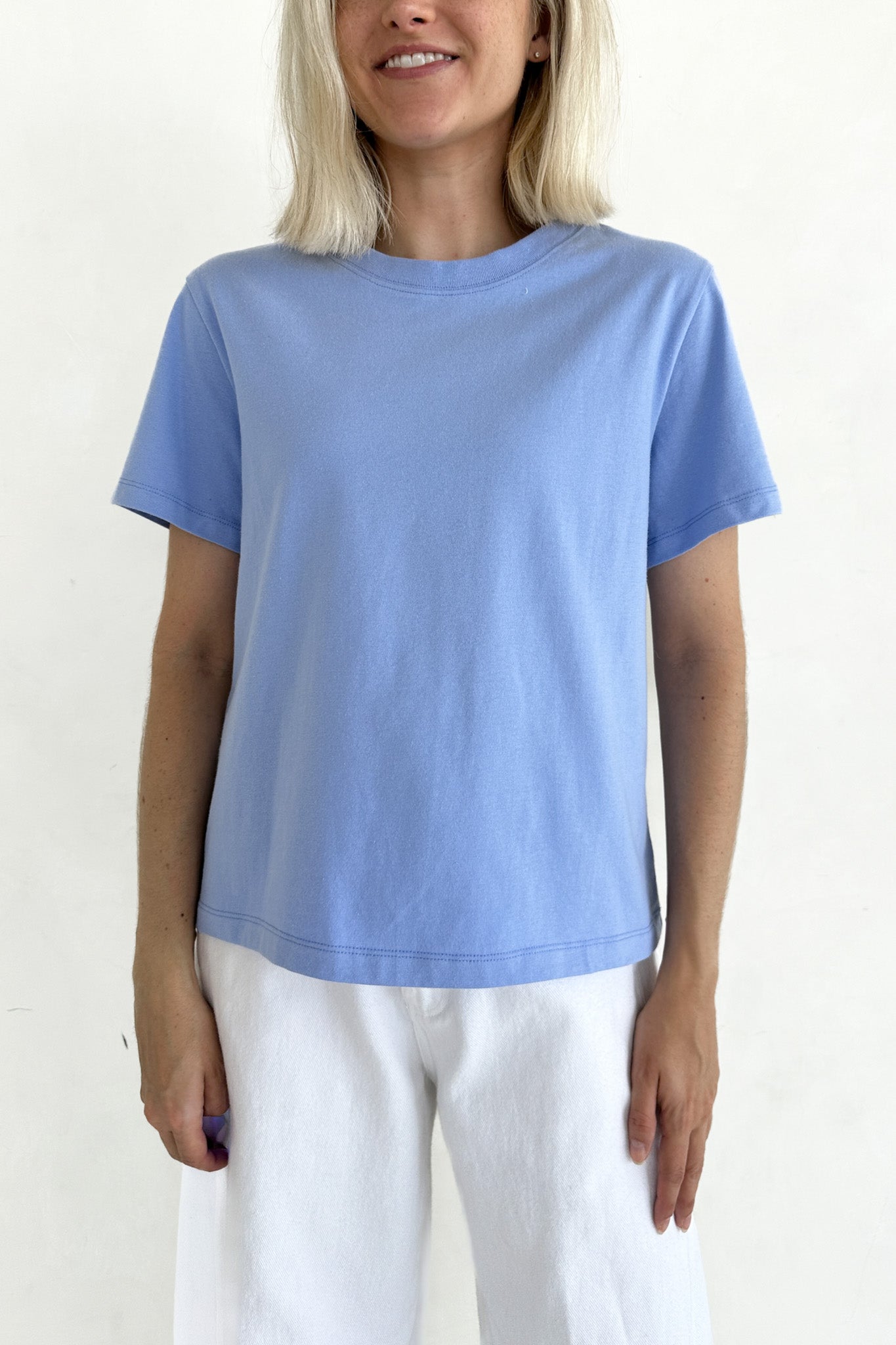 A woman wearing a light blue short-sleeve crewneck t-shirt and white pants, standing against a plain off-white background.
