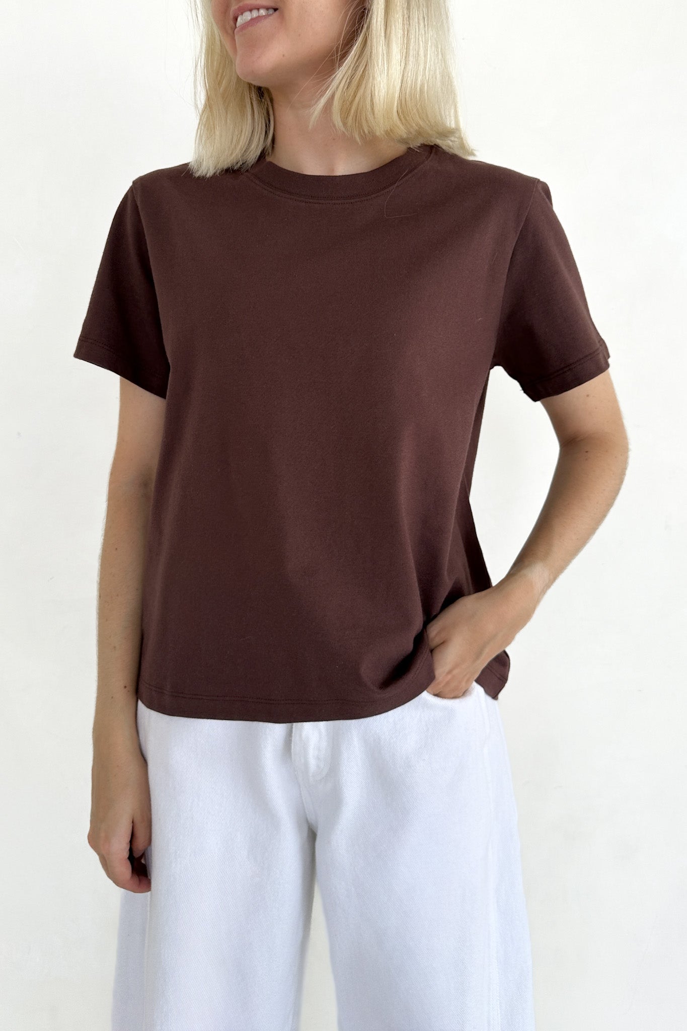 A woman wearing a plain brown short-sleeve crewneck t-shirt and white pants, shown from the chin down against a light off-white background.