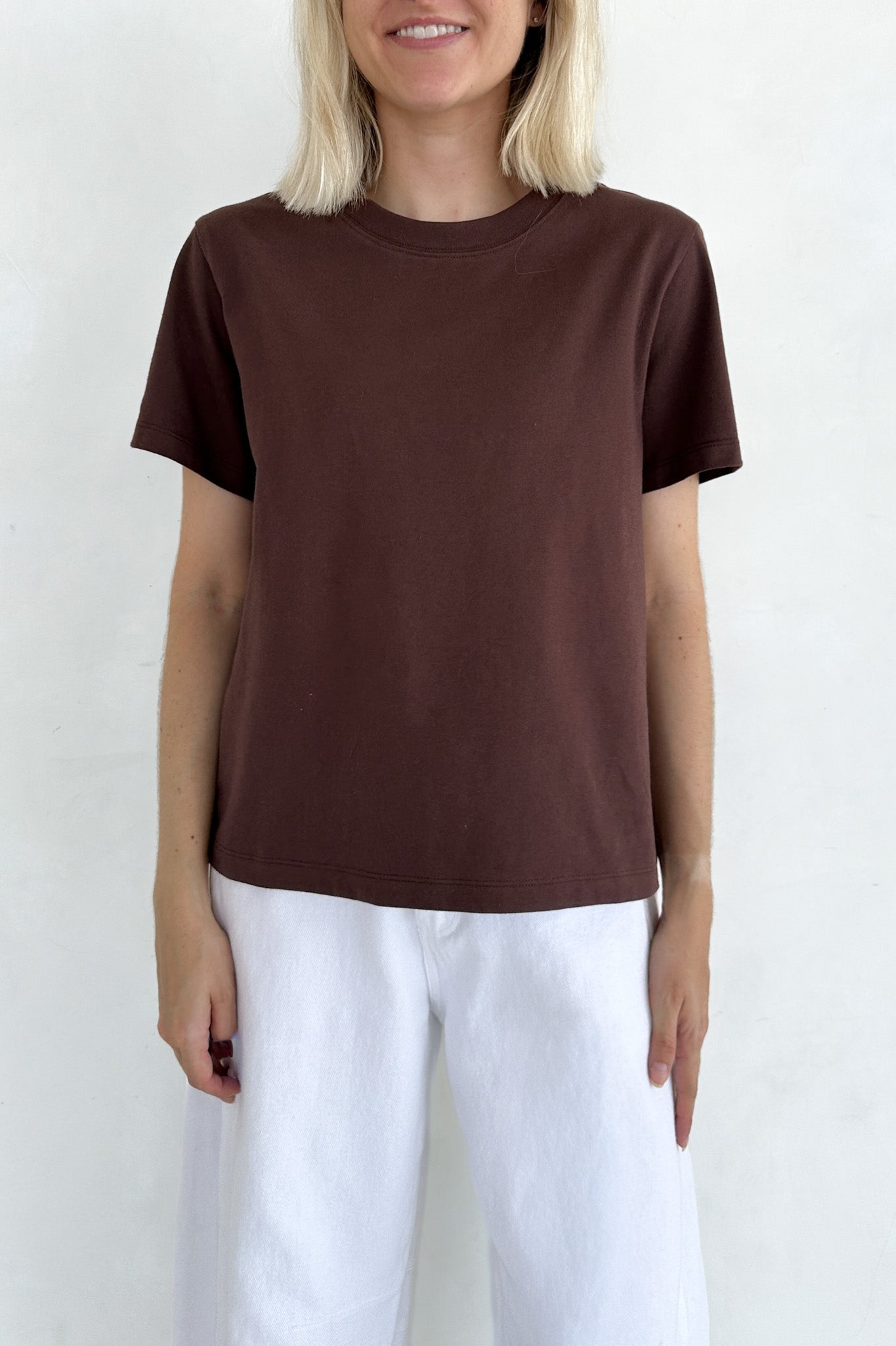 A woman wearing a dark brown short-sleeve crewneck t-shirt and white pants, standing against a plain white wall.