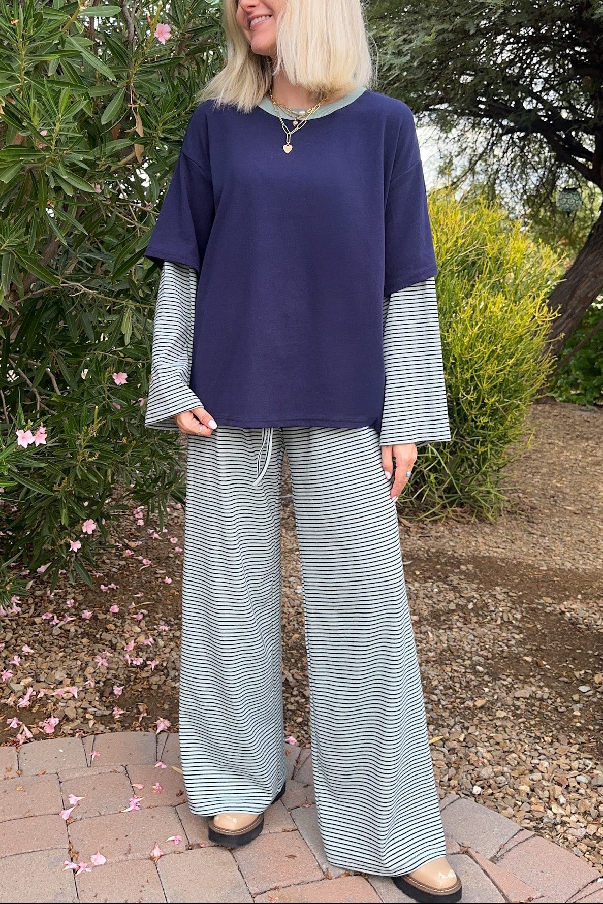 A woman stands outdoors in a garden wearing a navy blue top with long light blue and white striped sleeves and matching wide-leg striped pants.