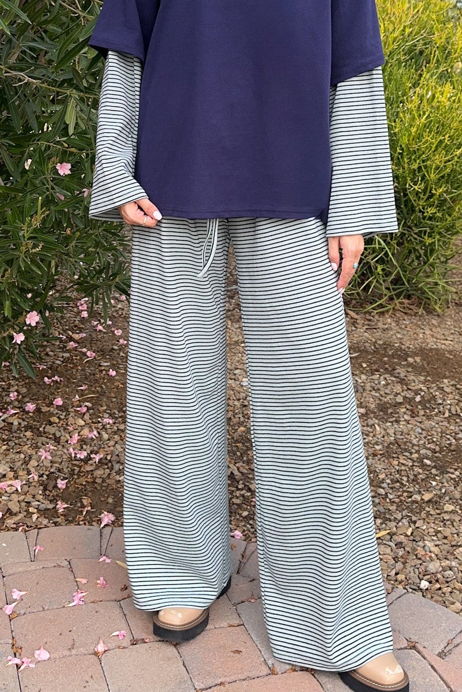 A woman stands outdoors in a garden wearing a navy blue top with long light blue and white striped sleeves and matching wide-leg striped pants.