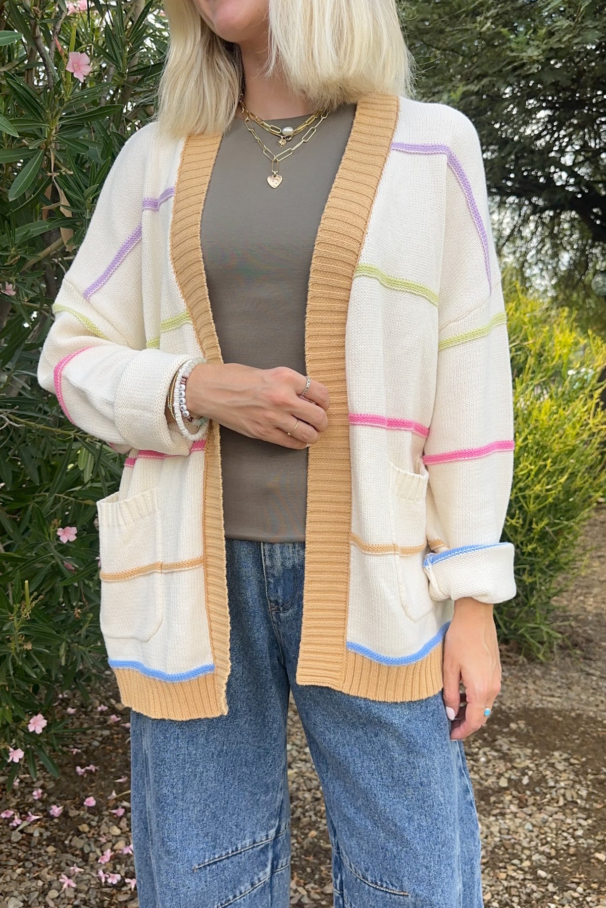 A woman wearing a cream-colored knit cardigan with pastel pink, purple, green, and blue stripes, styled over a taupe top and blue jeans in an outdoor garden setting.