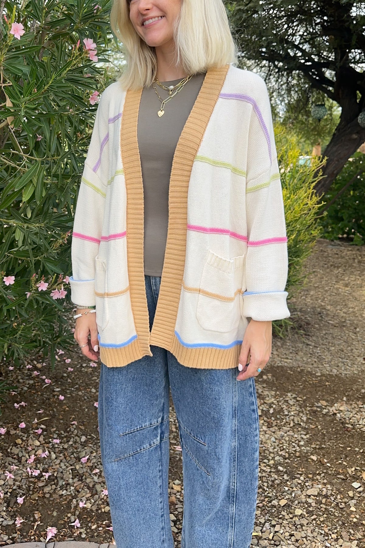 A woman wearing a cream-colored knit cardigan with pastel stripes in pink, purple, green, and blue, styled over a brown top and blue jeans in an outdoor garden setting.
