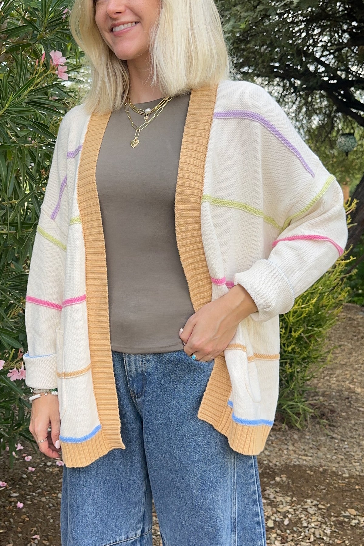 A woman wearing a cream open-front cardigan with mustard yellow ribbed trim and pastel pink, purple, green, and blue stripes, styled over a taupe top and blue jeans in an outdoor garden setting.