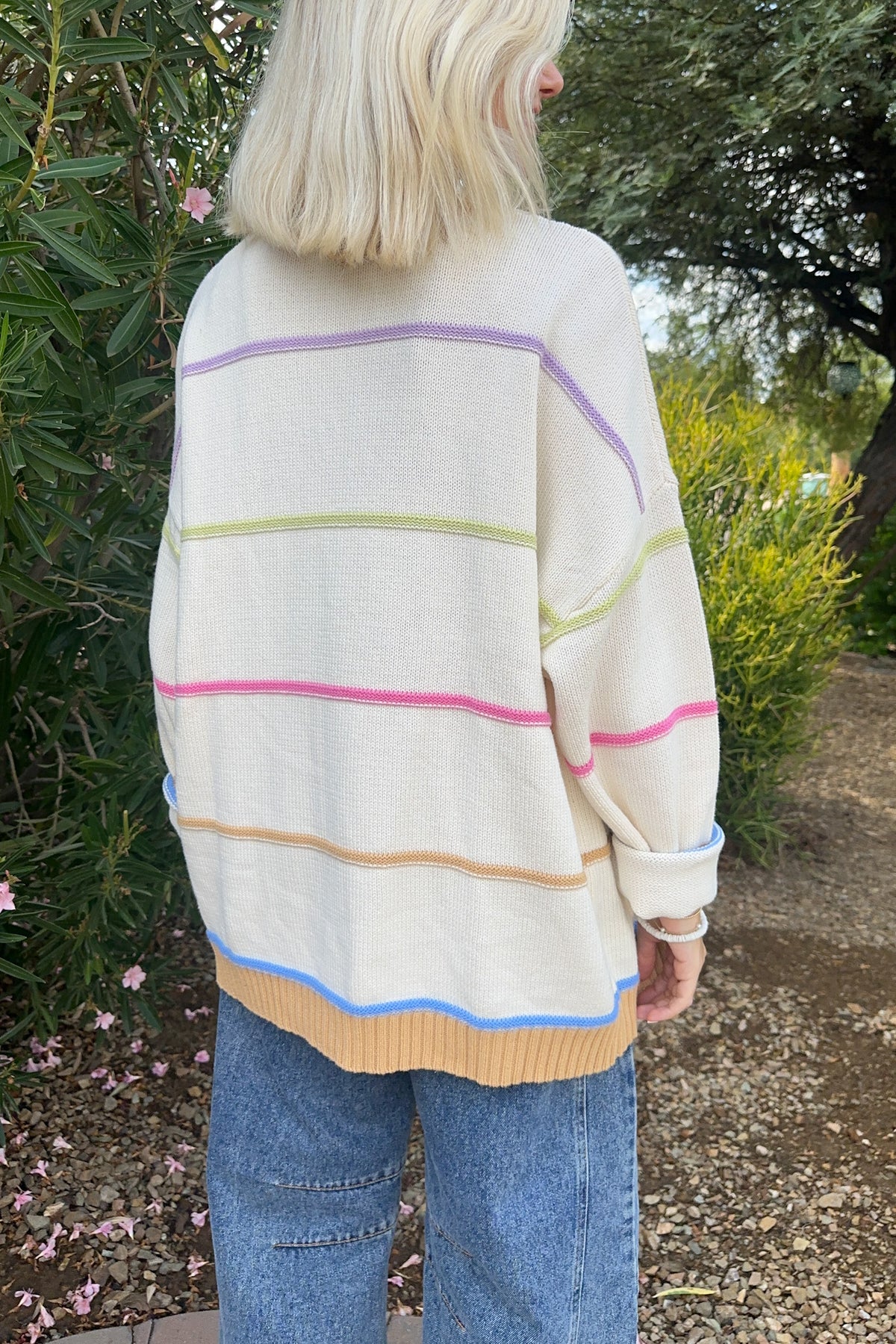 A person viewed from behind wearing an oversized cream knit sweater with thin horizontal pastel stripes in purple, green, pink, beige, and blue, paired with blue jeans in an outdoor garden setting.