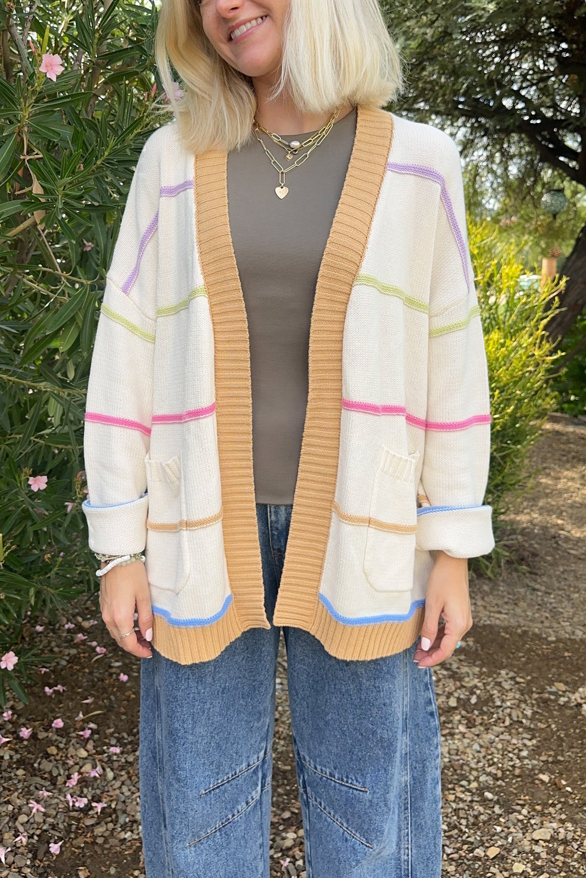 A woman wearing a cream-colored knit cardigan with pastel pink, green, and blue stripes, styled over a brown top and blue jeans in an outdoor garden setting.