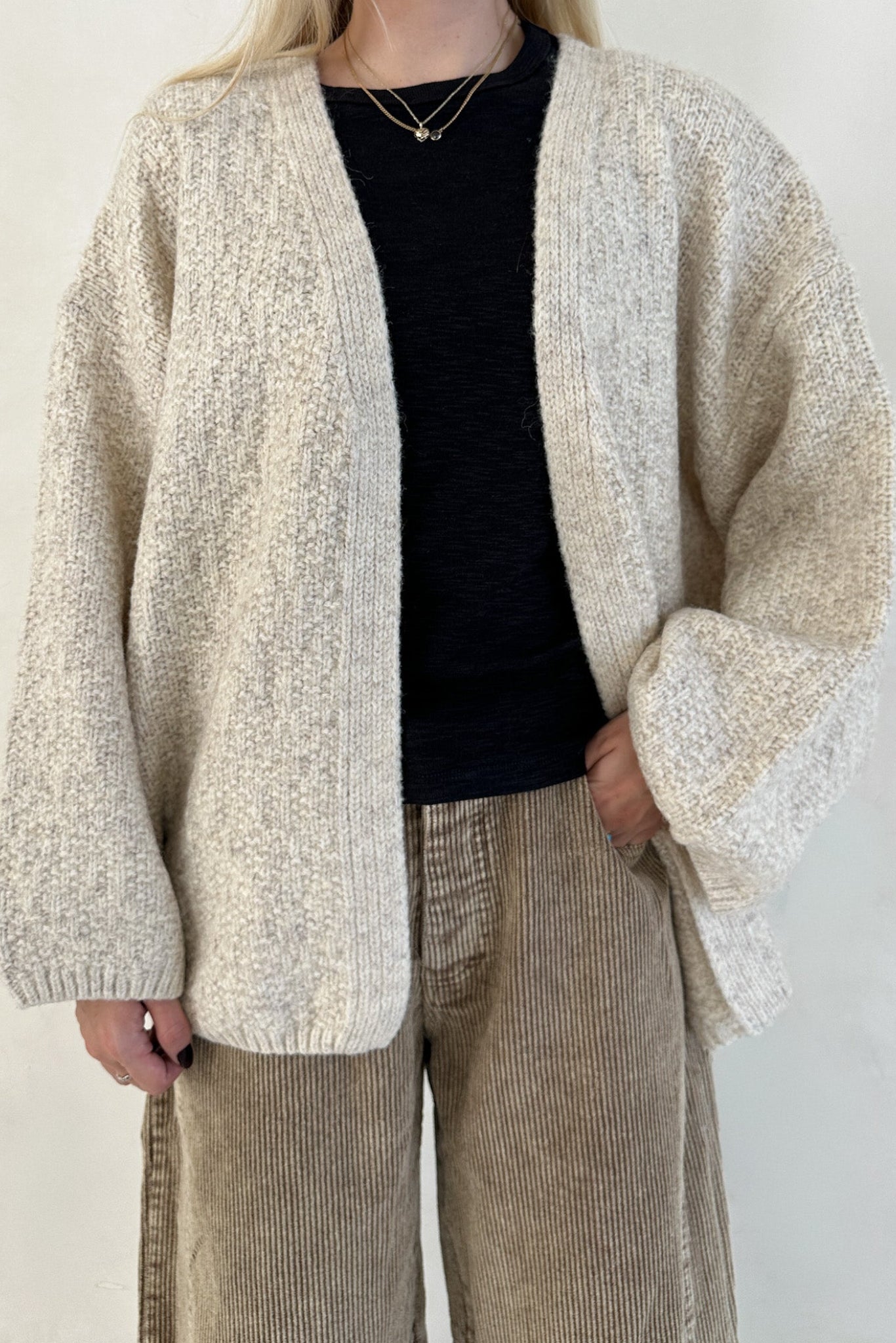 Person wearing a beige cardigan, black top, and brown pants against a white background