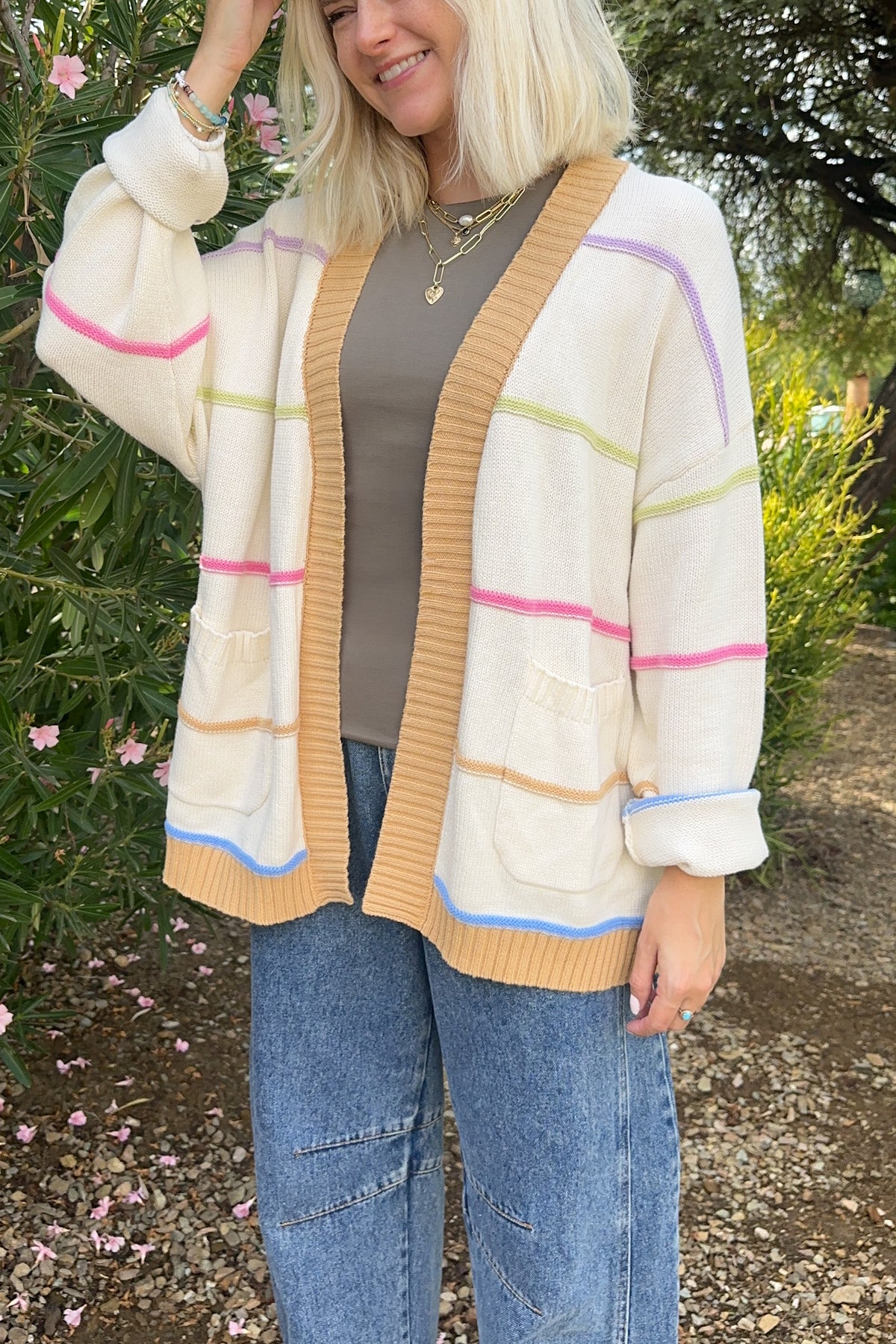 A woman wearing a cream-colored knit cardigan with pastel pink, green, and blue horizontal stripes, styled over a brown top and blue jeans in an outdoor garden setting.