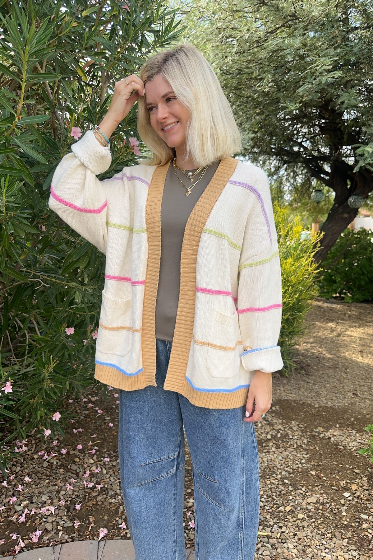 A woman with blonde hair stands outdoors wearing a cream-colored knit cardigan with pastel pink, green, and blue stripes, a ribbed mustard yellow trim, and a gray top tucked into blue jeans.