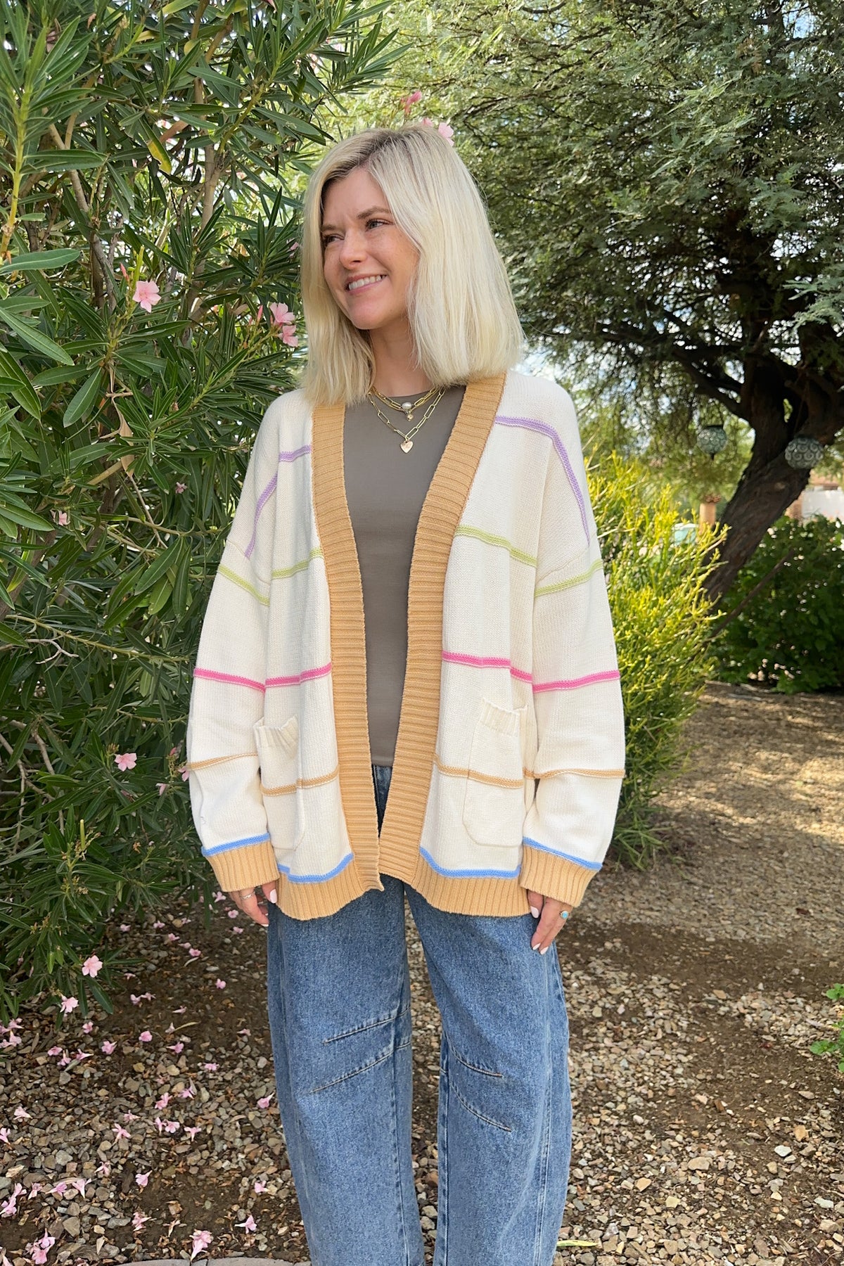 A woman with blonde hair stands outdoors in a garden wearing a cream-colored striped cardigan with mustard yellow ribbed trim, a gray top, and blue jeans.