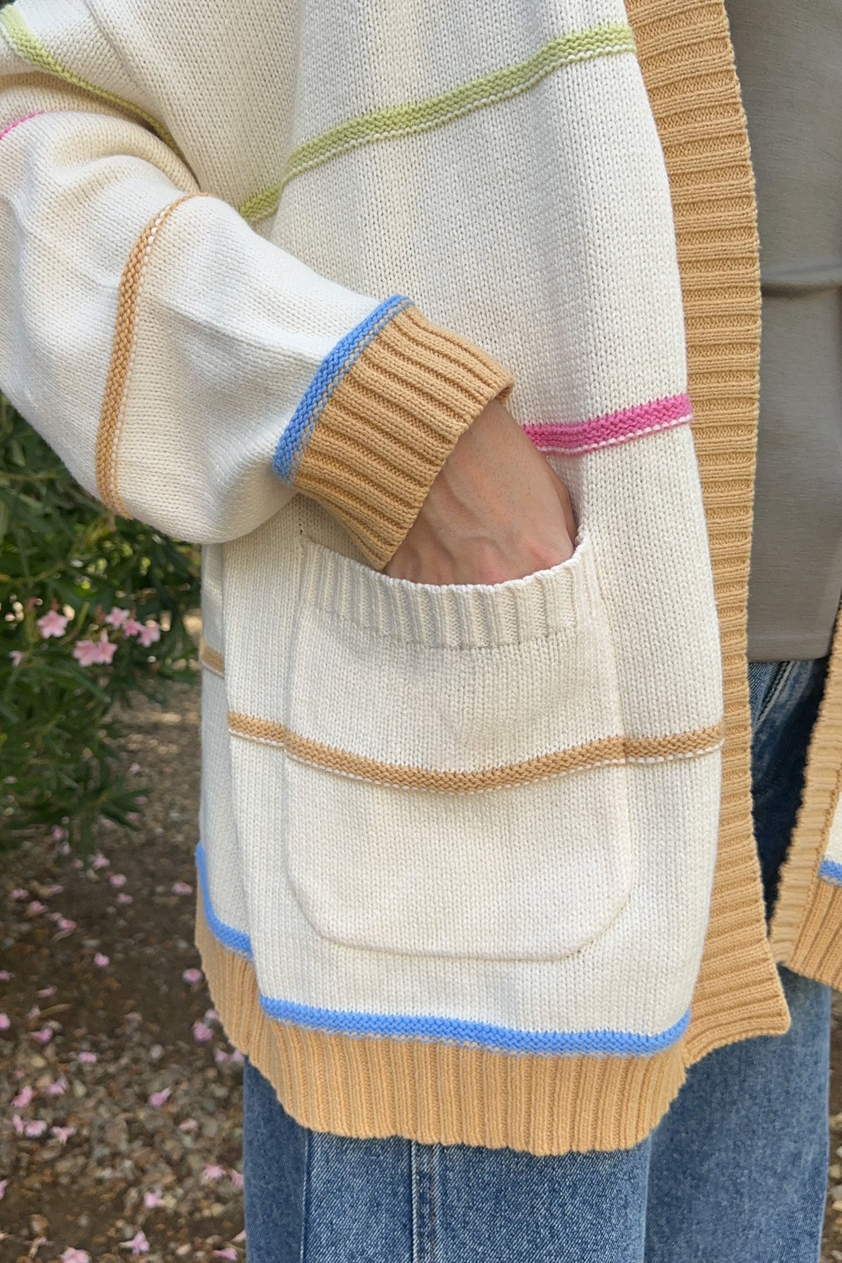 A close-up of a person wearing a cream knit cardigan with tan, light green, pink, and blue striped accents, featuring a front pocket and ribbed cuffs and hem, paired with blue jeans.