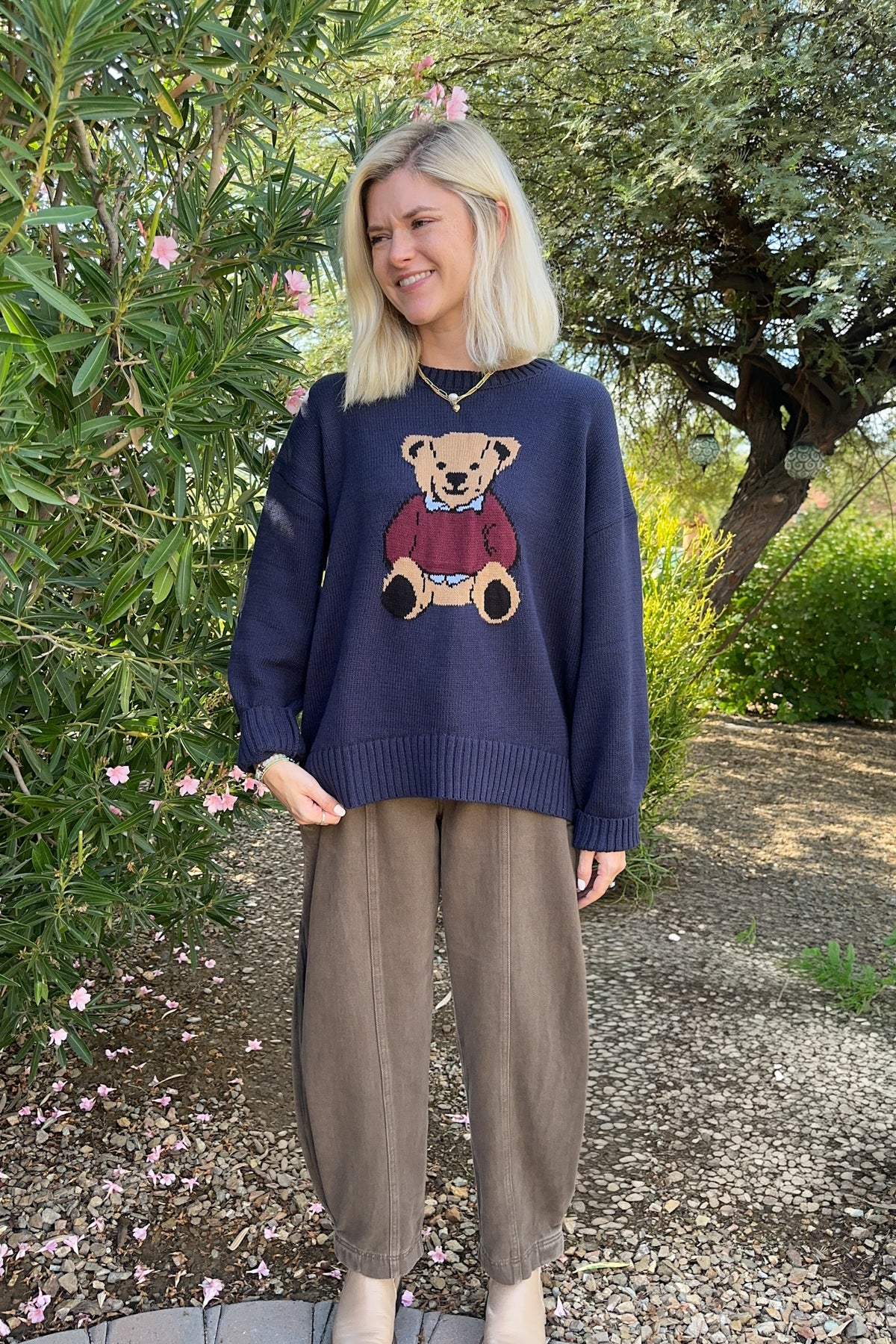 A woman wearing a navy blue crewneck sweater with a teddy bear graphic and brown wide-leg pants, standing outdoors in front of green trees and bushes.