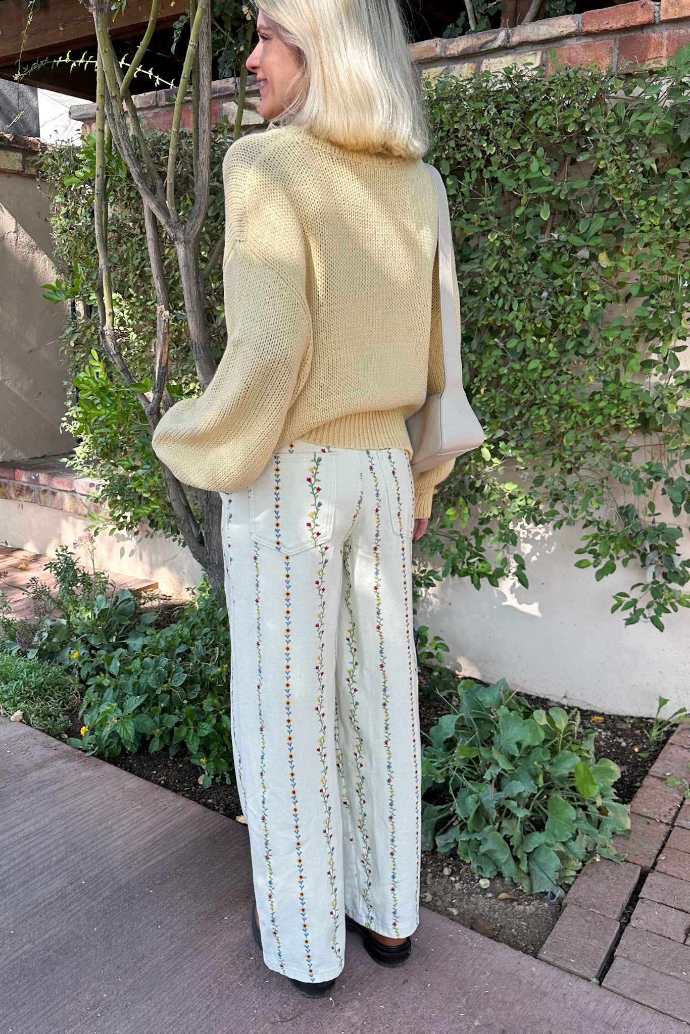 Woman in a yellow sweater and white pants standing outdoors with greenery in the background