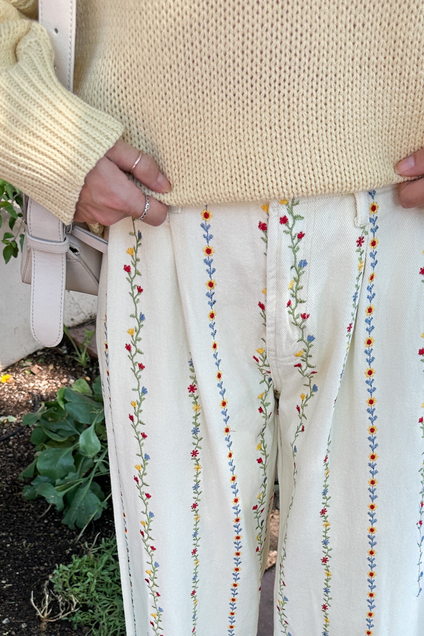 Person wearing a beige sweater and white pants with colorful embroidery, standing outdoors.
