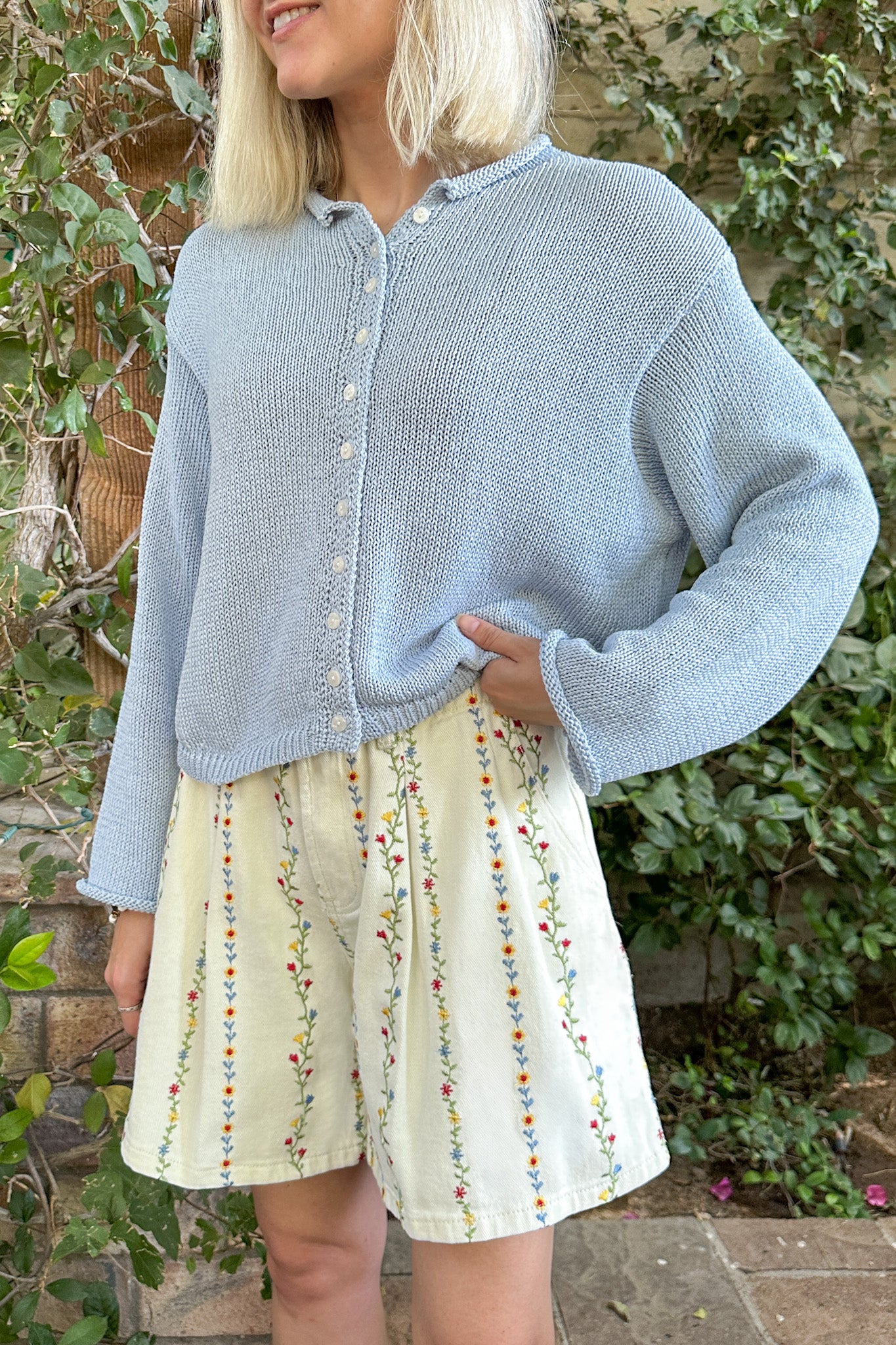 Woman wearing a light blue cardigan over a patterned skirt with a natural background