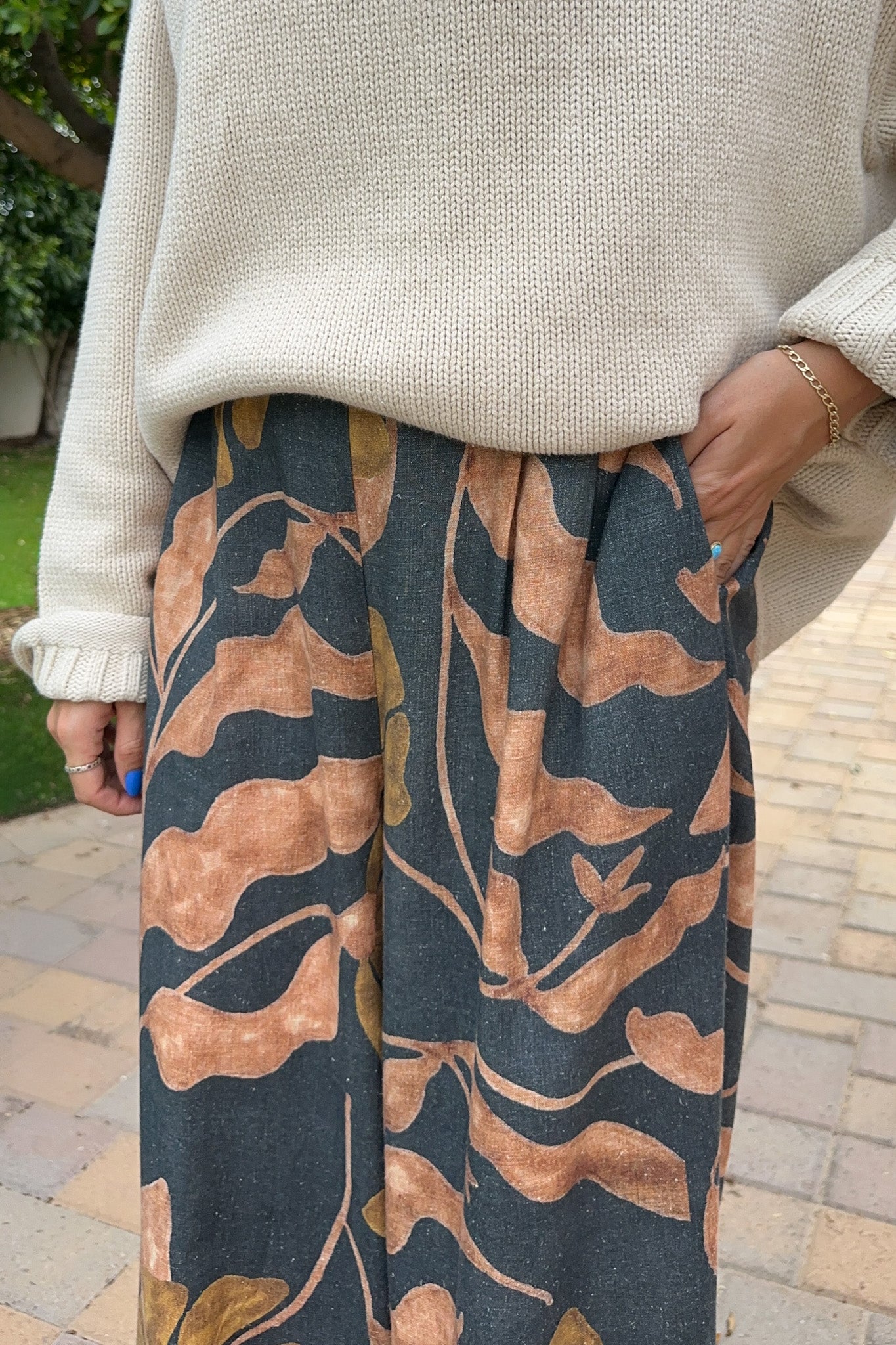 A person wearing a cream knit sweater and wide-leg pleated trousers with a dark charcoal and tan botanical leaf pattern, standing outdoors on a paved walkway.