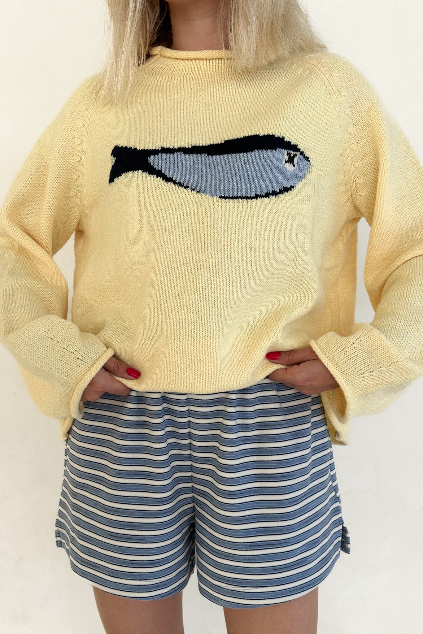 Person wearing a yellow sweater with a fish design and blue striped shorts on a plain background
