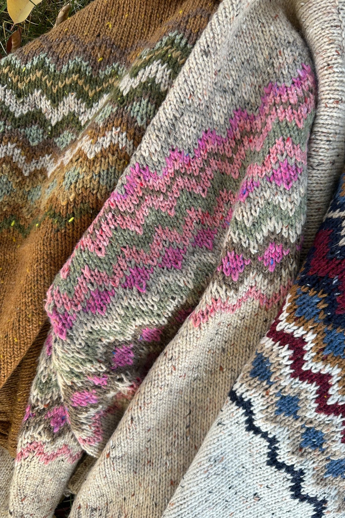Close-up of knitted wool garments with Fair Isle patterns in pink, blue, brown, green, and cream, draped together outdoors.
