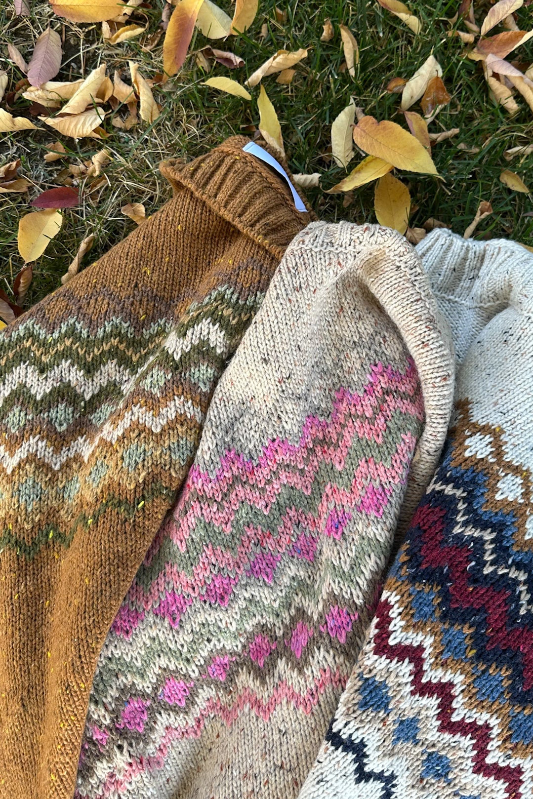 Three hand-knit sweaters with colorful Fair Isle patterns are laid out on grass covered with fallen autumn leaves. The sweaters feature different colorways: one brown and mustard, one cream and pink, and one cream and burgundy.