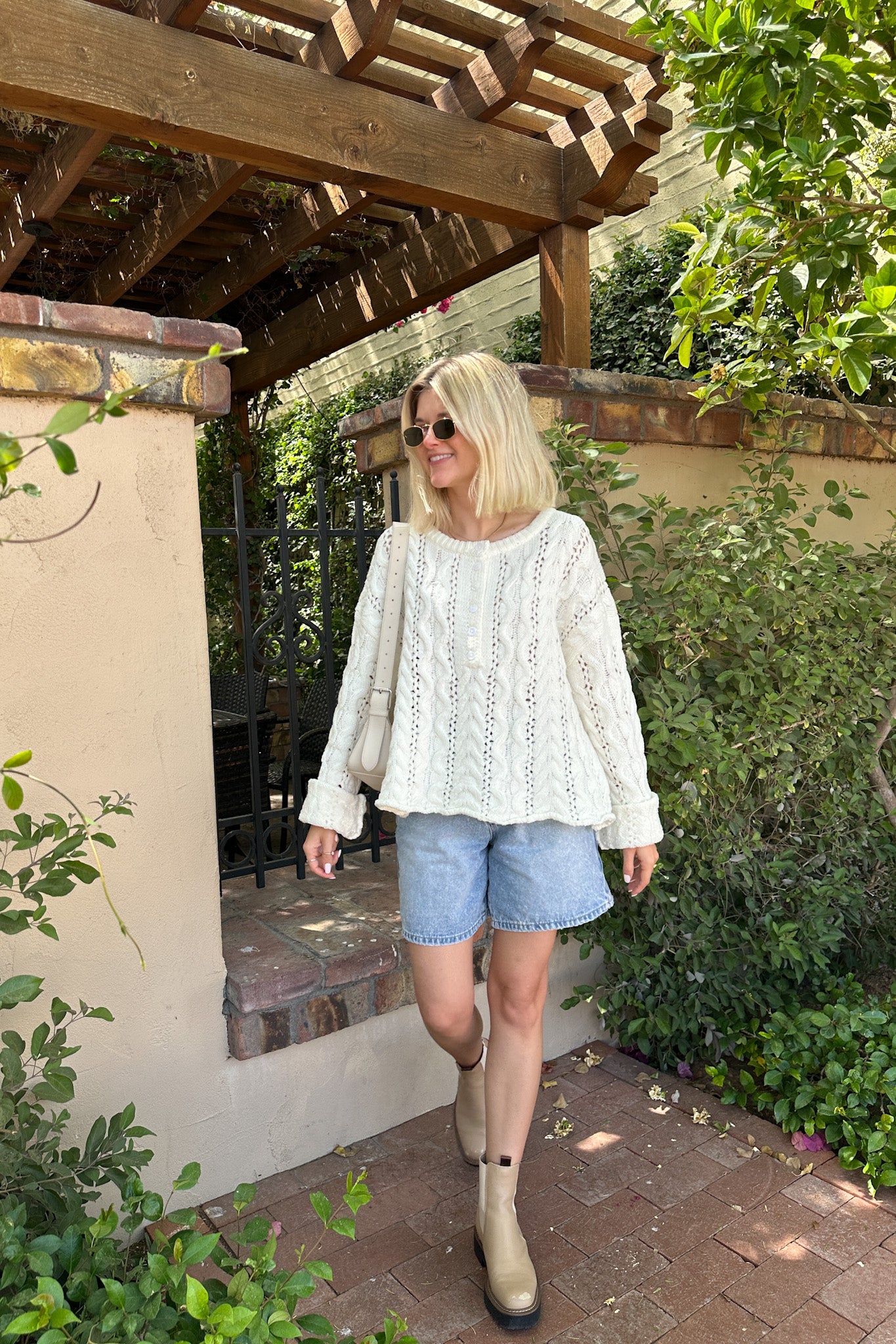 Woman in a white lace top and denim shorts walking outdoors under a wooden pergola.