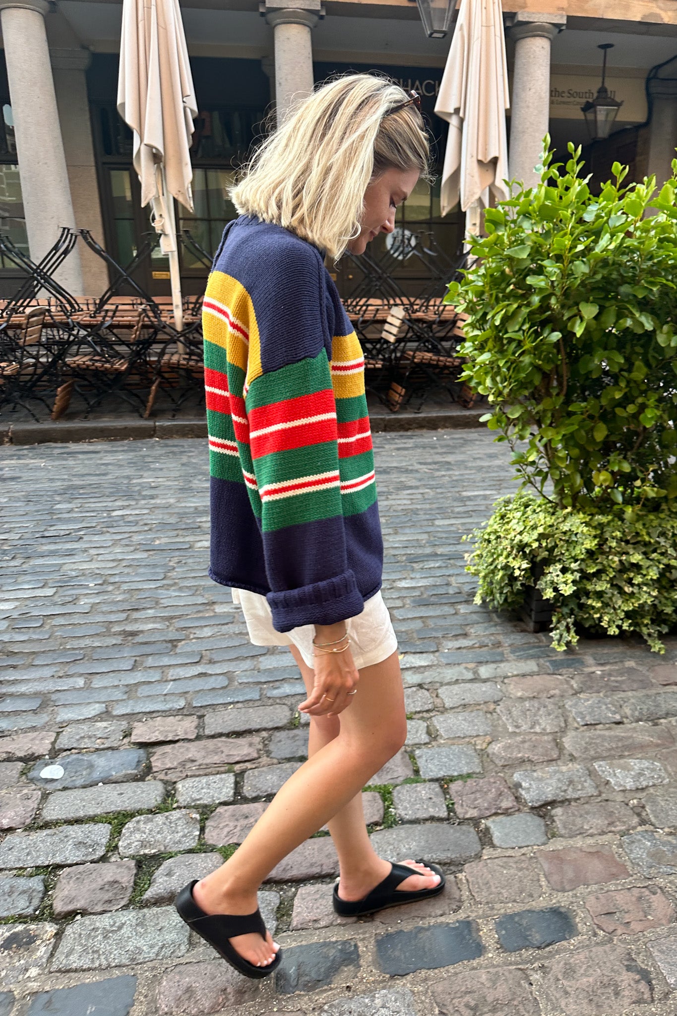A woman wearing a colorful striped knit sweater, white shorts, and black sandals walking on a cobblestone street in front of an outdoor cafe with closed umbrellas.