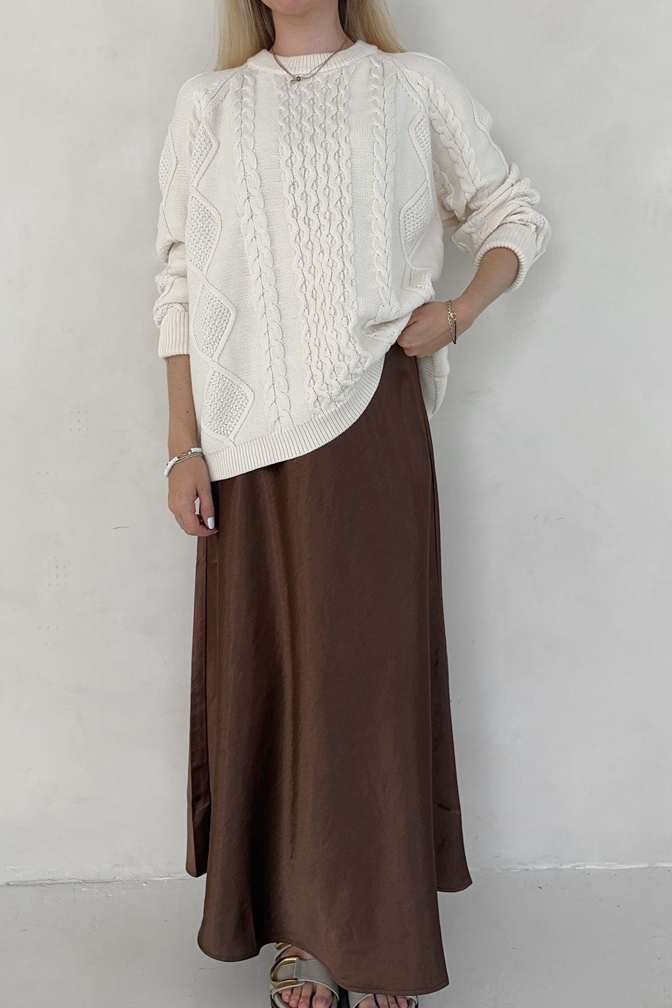 Person wearing a white cable knit sweater and brown skirt against a plain background