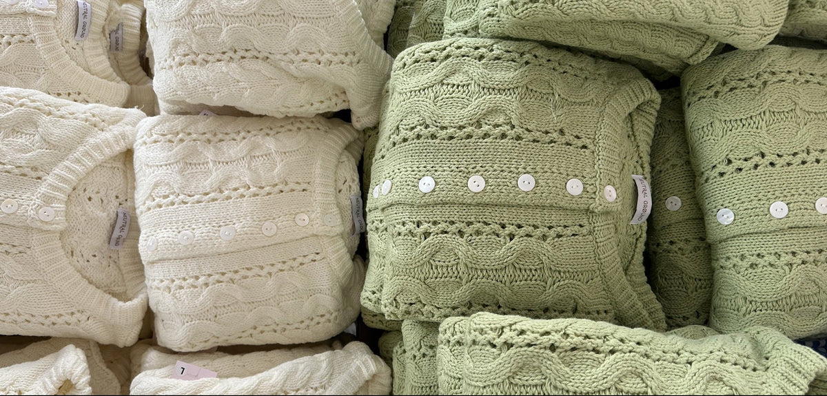 A close-up of folded cable-knit sweaters stacked together, showing cream and pale sage green colors with white decorative buttons.