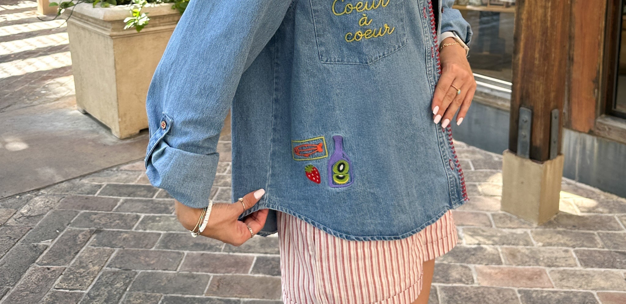 Person wearing a denim jacket with colorful patches on a paved outdoor area.