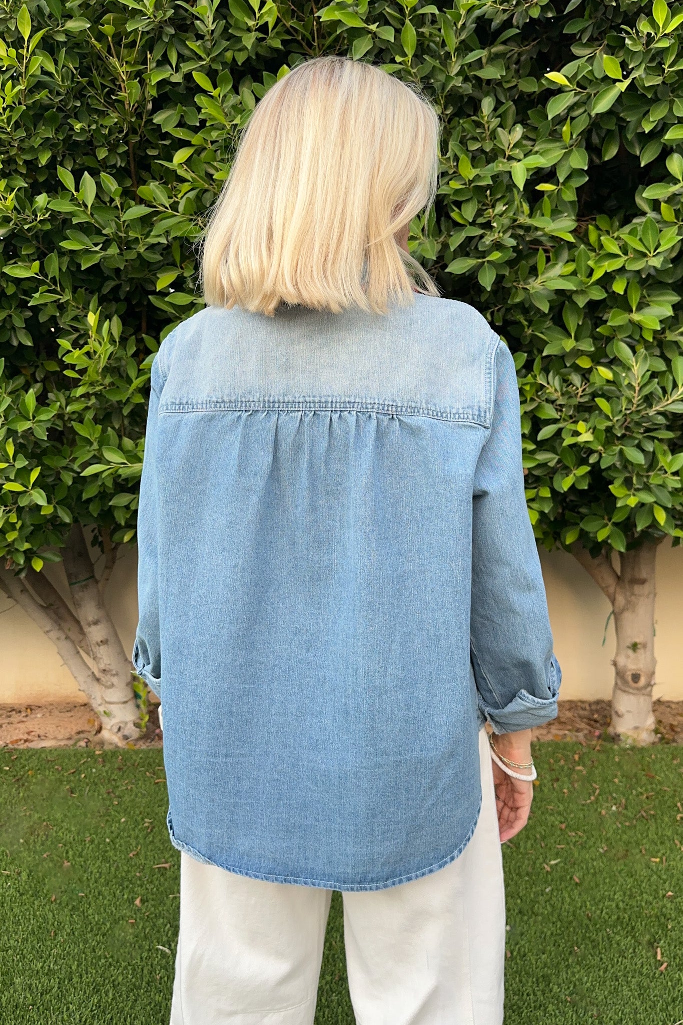 Person wearing a blue denim shirt and white pants standing in front of green bushes.
