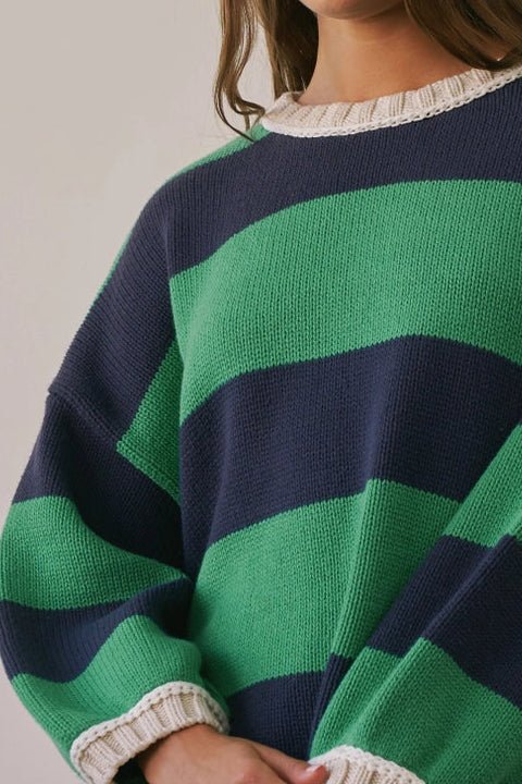 A woman wearing an oversized knit sweater with bold green and navy horizontal stripes, cream ribbed trim, and white shorts, photographed against a plain light gray studio background.